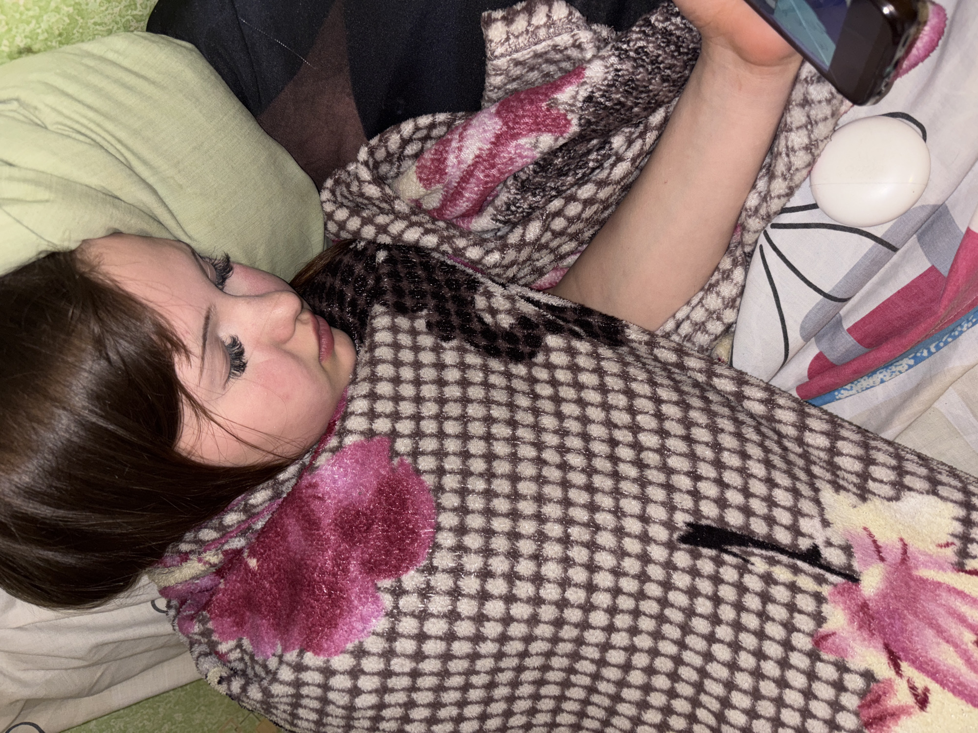 A young person lies on a bed, wrapped in a blanket with a brown and beige checkered pattern interspersed with pink floral designs. The person's head rests on a light green pillow. They are holding a smartphone in their right hand, and a white circular object, possibly an earbud case, rests next to the phone. The bedsheets are patterned with stripes and pink floral motifs. The scene is dimly lit, with the focus being on the person's face and the surrounding bedding. The location information is irrelevant to the visual content of the image.