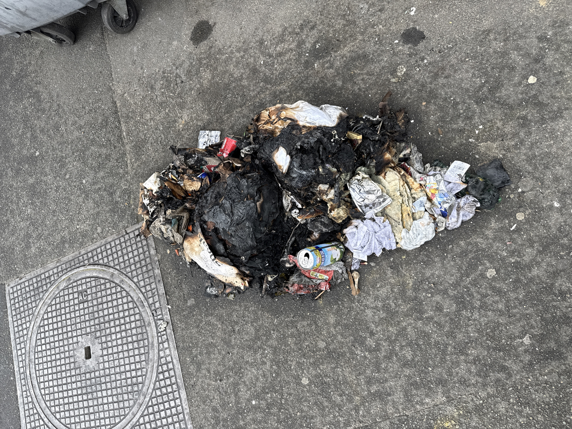The image shows a pile of burnt trash on a concrete surface next to a manhole cover and a partially visible garbage container with wheels. The trash includes charred material, paper, cans, and clothing remnants. The location is implied to be Sarcelles, France. There are no people visible in the frame. The main subject is the trash pile and its location.