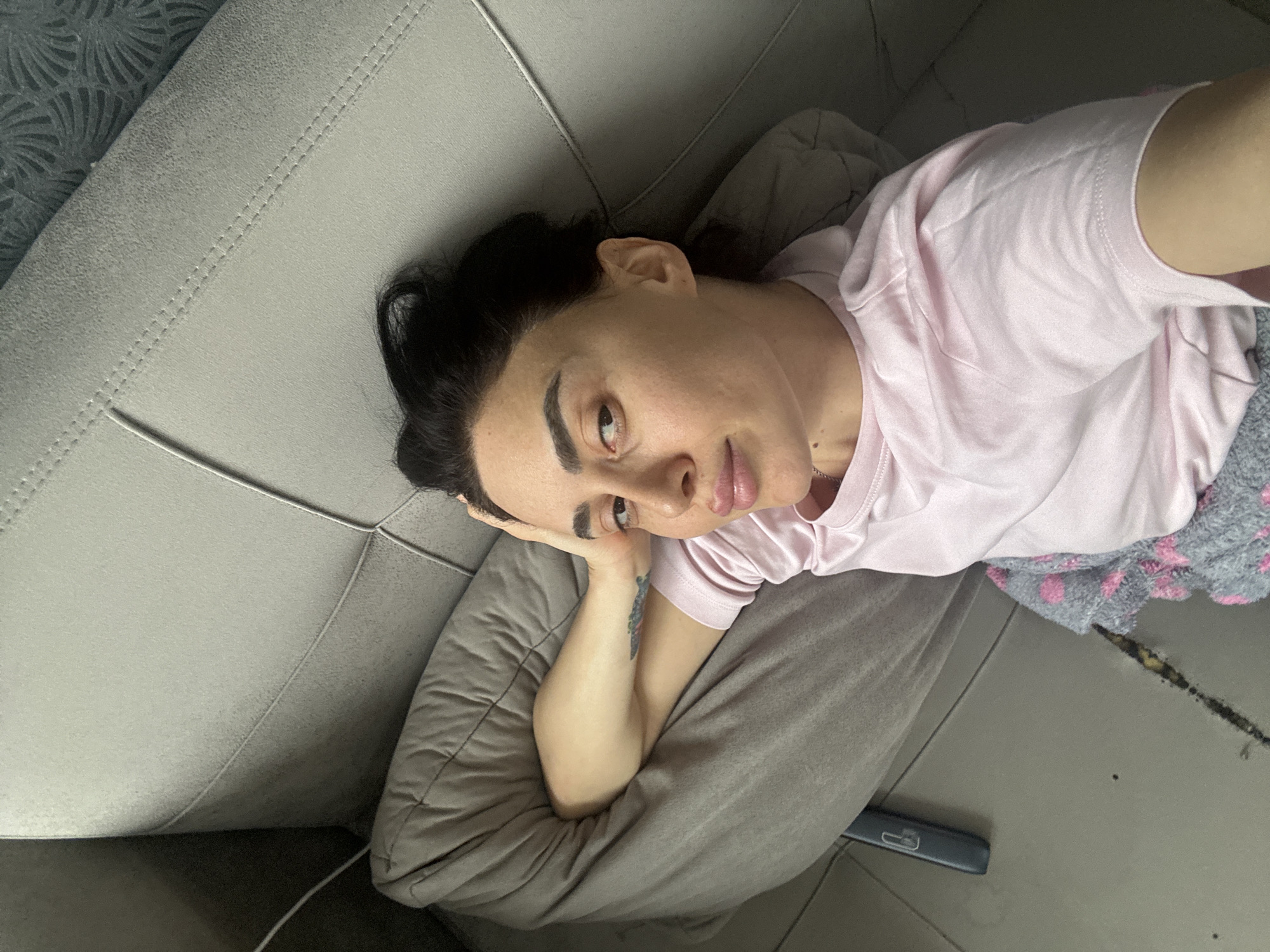 A person is seen lying down on a light grey sofa in an indoor setting, likely a living room or bedroom in Tbilisi, Georgia. The individual, appearing to be a woman, has dark hair pulled back and defined eyebrows. She is wearing a light pink short-sleeved t-shirt. Her head is propped up on a grey pillow, and she is looking directly at the camera with a neutral, slightly relaxed expression. Her lips are slightly parted. A tattoo is visible on the inner side of her left forearm.
The sofa features visible stitching details. To the right of the person, a grey blanket with pink polka dots is partially visible. The floor is dark grey or black, and a notable dark, elongated stain or mark is present on it near the blanket. A dark blue remote control is also visible on the floor. In the upper left background, a wall with a decorative, fan-like patterned wallpaper can be seen. The lighting suggests it is daytime. There is no visible text in the image.