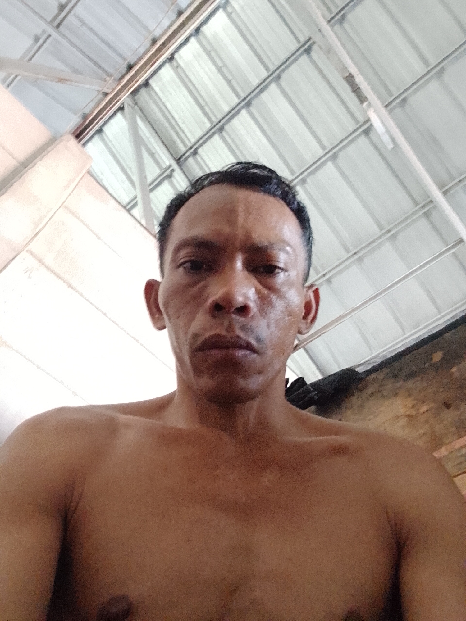 The image shows a shirtless middle-aged man with dark, receding hair, standing indoors in Bukit Ketok, Indonesia. He is facing the camera, appearing to take a selfie. The background includes an exposed metal roof structure. The man is the primary subject of the image. There is no visible interaction or action taking place. The environment appears to be a simple indoor space.