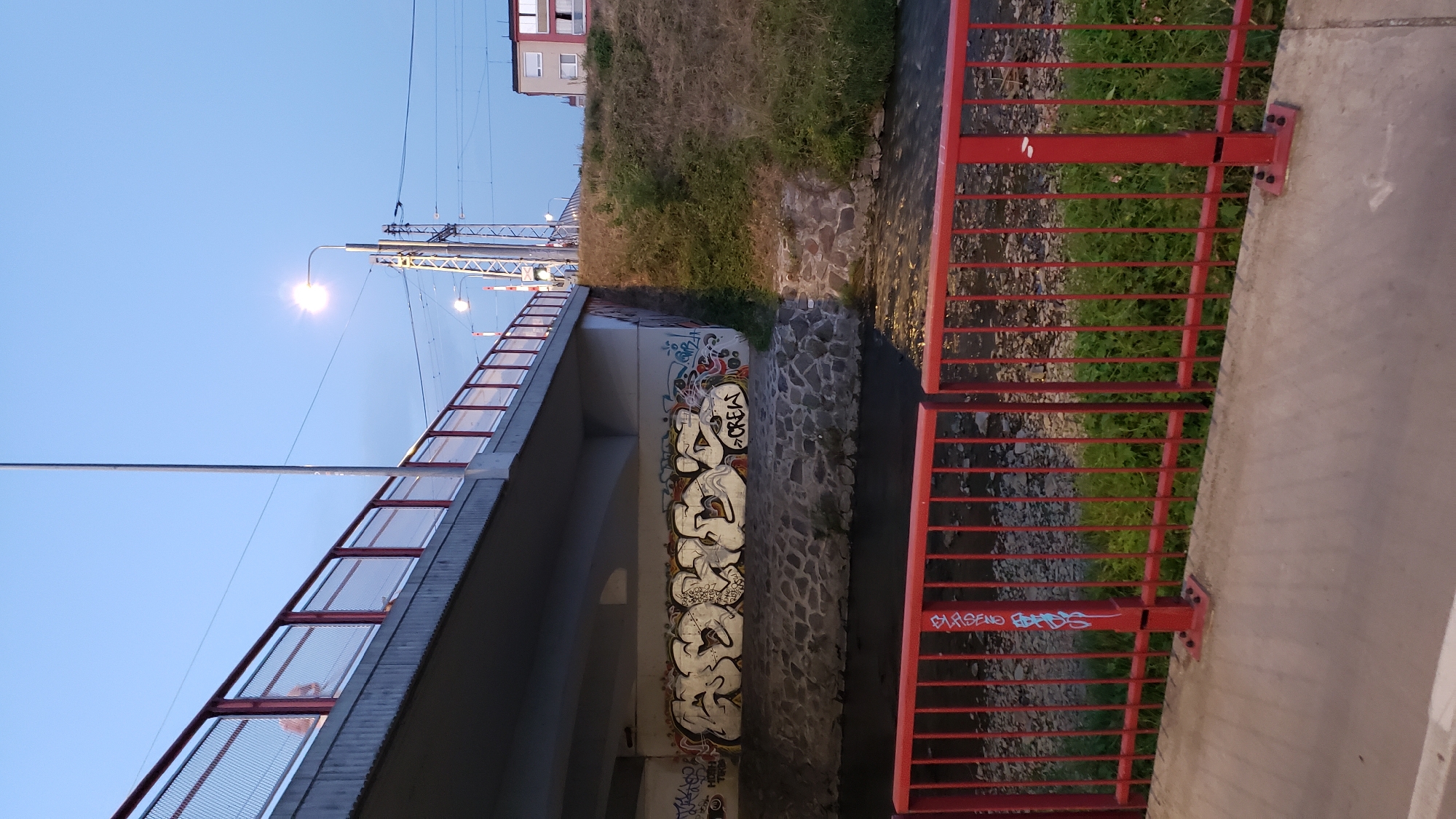 The image captures a view under a concrete bridge, likely in the city of Prešov, Slovakia, given the context of the request. The bridge spans a narrow, dark body of water, which appears to be a stream or small river, with a stone embankment visible along its edge. The underside of the bridge is adorned with graffiti, predominantly in white and black, featuring stylized lettering. Notably, the words "BORS CREW" are clearly visible.
To the right of the bridge, a grassy slope rises, covered with dry grass and some green vegetation. A streetlamp illuminates the scene from the left, casting a glow on the graffiti. In the background, power lines and utility poles suggest an urban or semi-urban setting. A modern, multi-story building with white window frames is visible in the far right background.
In the foreground, a bright red metal railing separates the viewer from the water and the bridge's embankment. The railing has vertical bars and horizontal supports, with some minor graffiti tags in blue and red visible on its posts. The ground in the immediate foreground is a concrete surface. The sky above is a pale blue, indicating either dawn or dusk. There are no visible people in the frame.