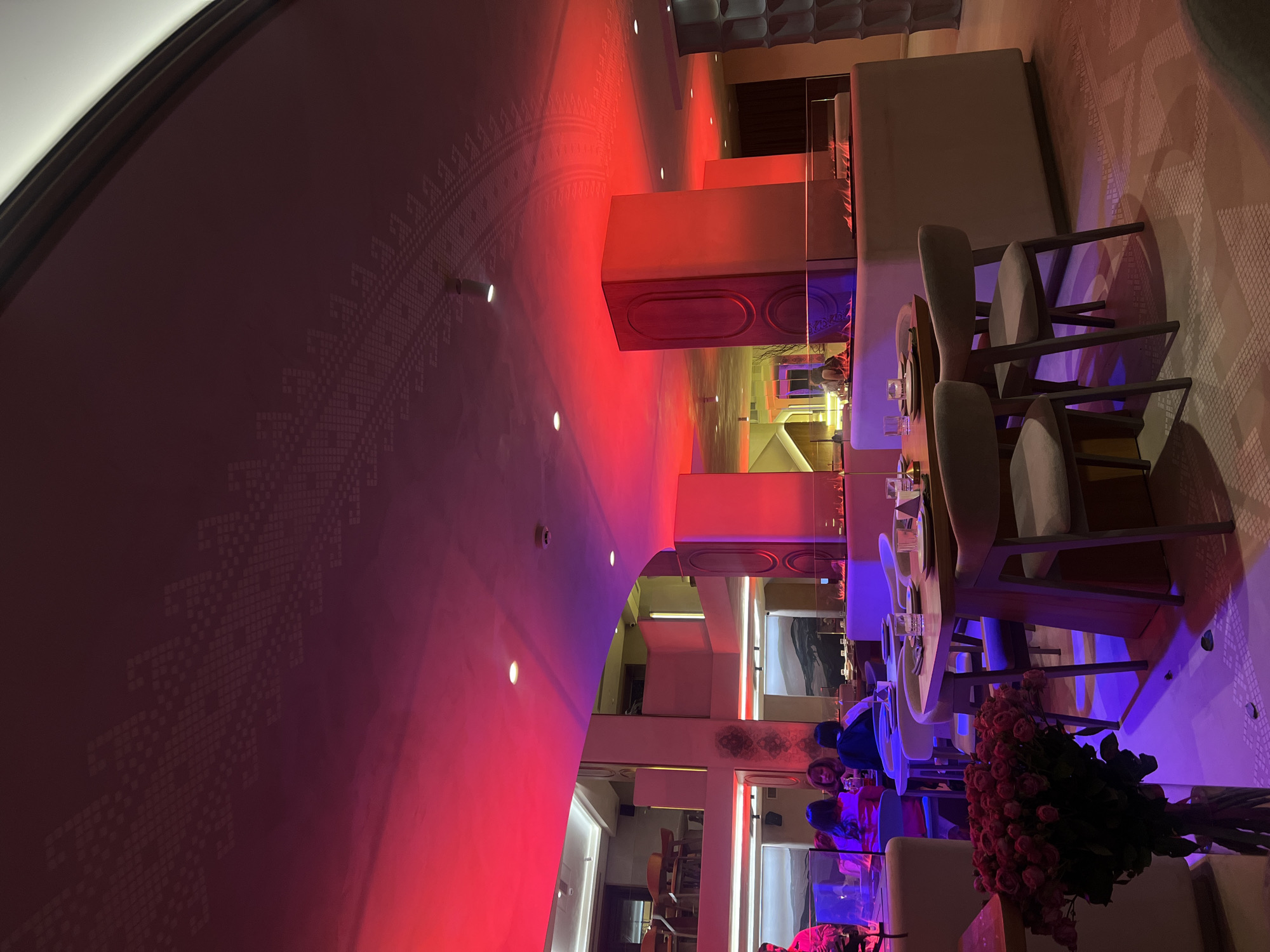 The image shows the interior of a restaurant with modern decor, bathed in red and purple lighting. Tables are set with glassware and plates, suggesting a dining setting. Seated individuals are visible, appearing to be engaged in a meal. Floral arrangements, specifically roses, adorn the foreground. Architectural features such as the arched ceiling and paneled walls contribute to the restaurant's ambiance. Given the location cues, this scene is likely situated in Yerevan, Armenia.