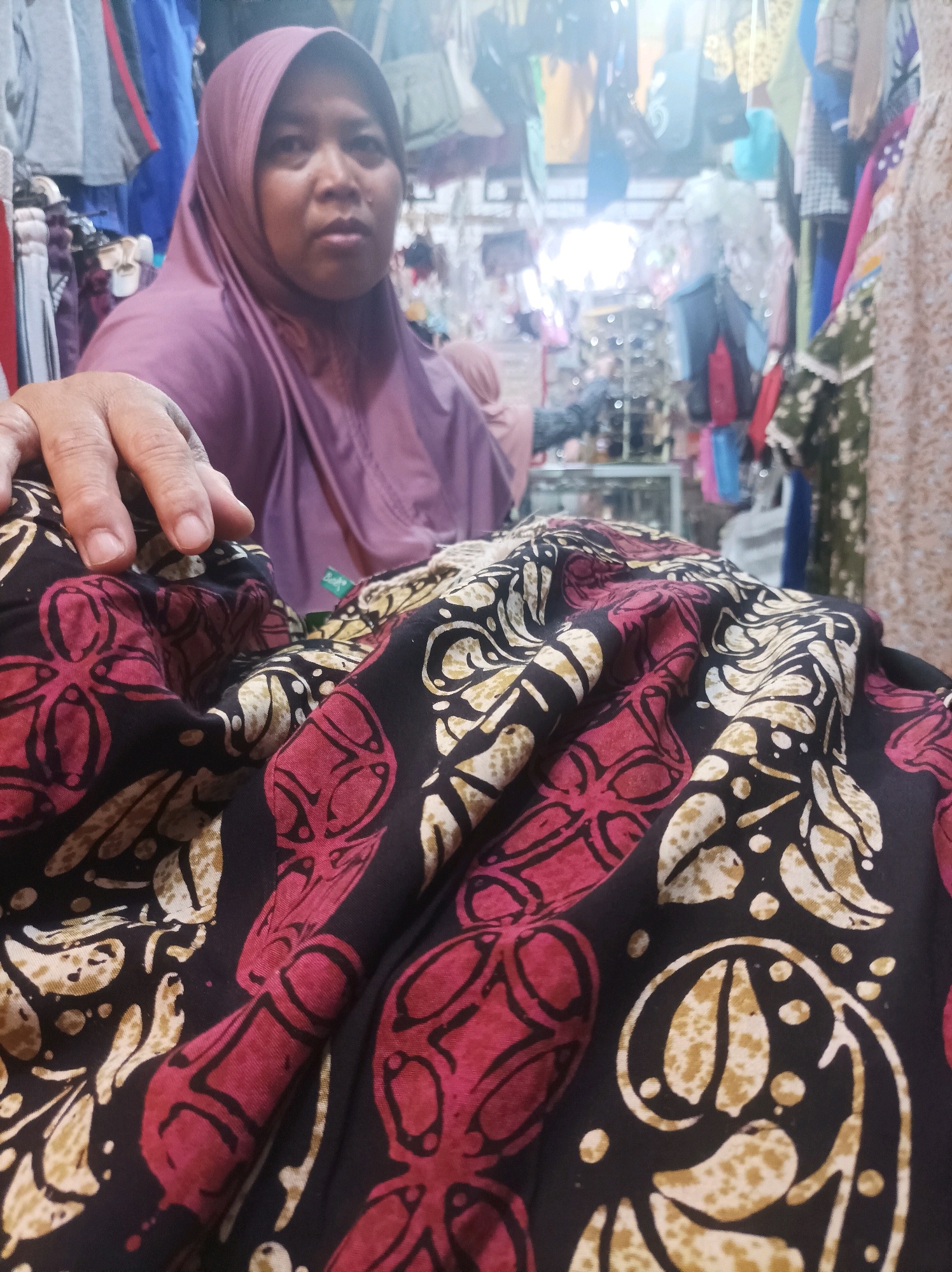 An Indonesian woman, wearing a purple hijab, is shown in a dimly lit shop, likely a clothing store. Her left hand is resting on a piece of fabric with a dark background and a bold pattern of red and cream-colored floral or organic shapes. The fabric is draped in the foreground, obscuring the lower part of her body and the rest of the shop's merchandise. The background is cluttered with various items of clothing and bags hanging on display racks, suggesting a retail environment. The overall lighting is soft and somewhat diffused, with a bright light source visible in the upper right quadrant of the image. The setting appears to be a market or a small shop in Sumuragung, Indonesia, given the cultural context implied by the hijab and the style of the merchandise.