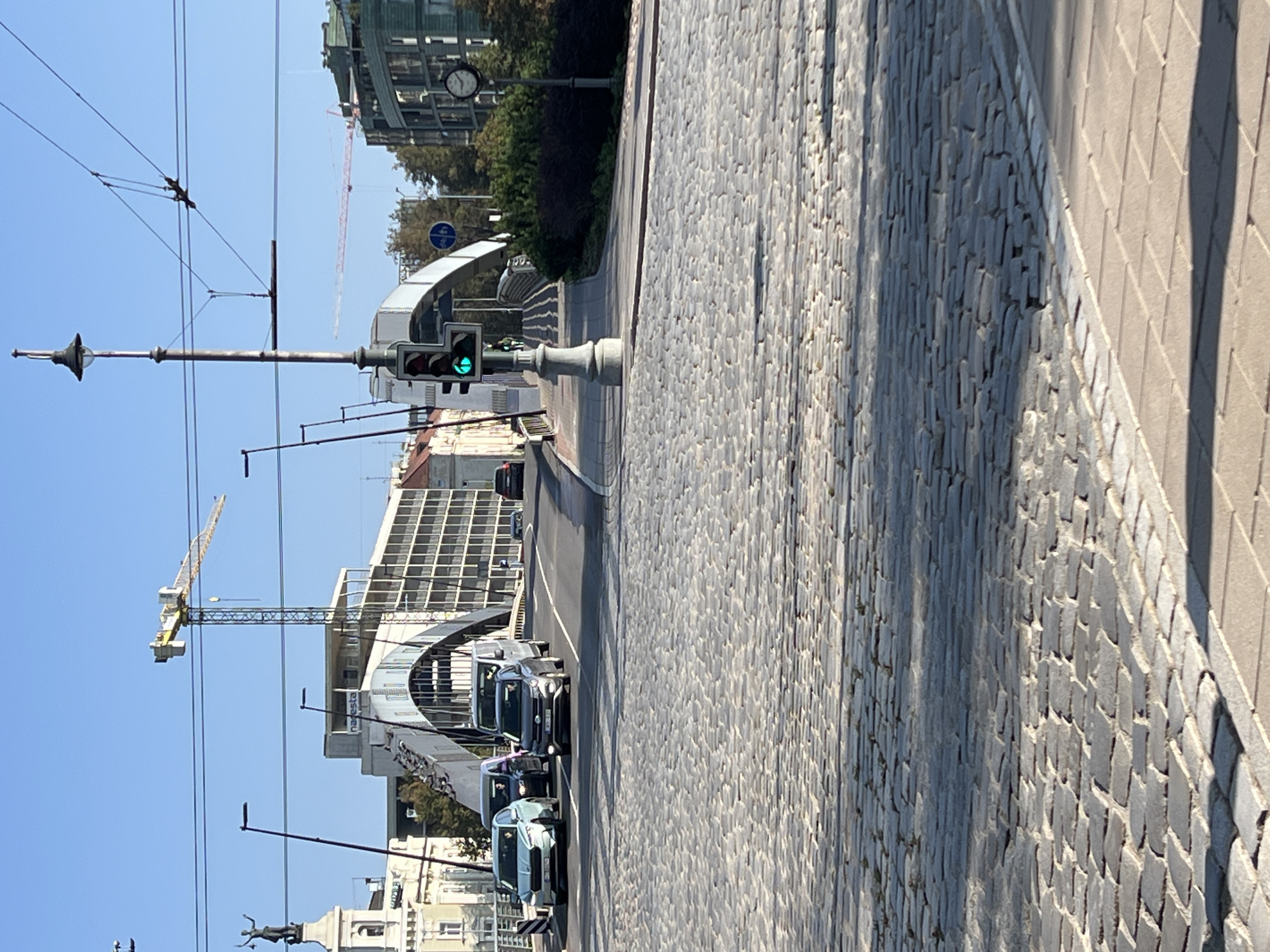 The image depicts a street scene in Vilnius, Lithuania, focusing on a bridge with a distinctive arched structure. In the foreground, a cobblestone street leads towards the bridge, with several vehicles, including cars and vans, traversing it. A traffic light, illuminated with a green signal, is visible on the left side of the street. In the background, a construction crane is prominent, suggesting ongoing development in the area. The architecture of the buildings lining the street suggests a historical European city. The sky is clear and blue, indicating fair weather.