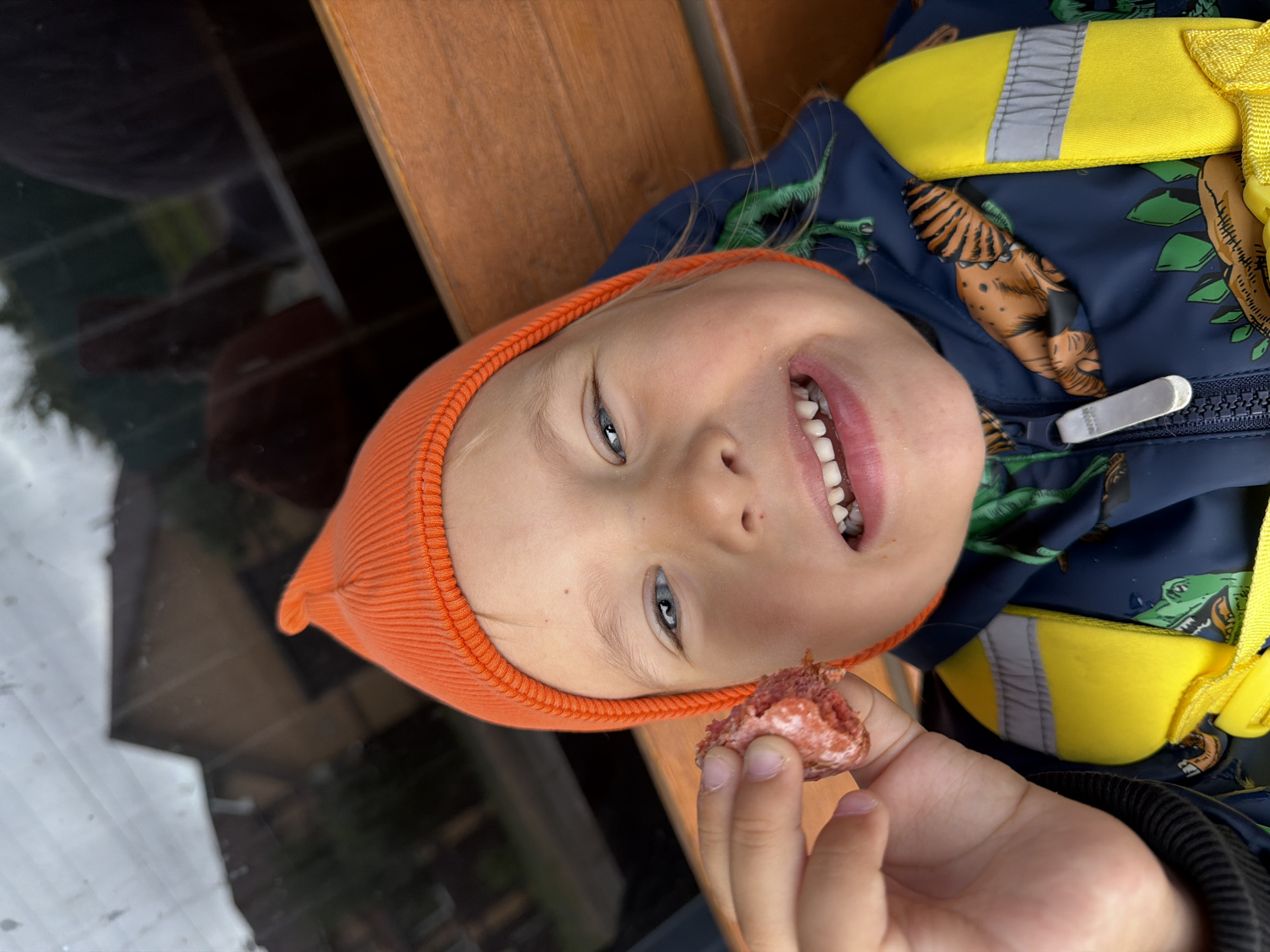 A close-up, eye-level, vertical shot shows a young child wearing an orange beanie and a dark blue jacket with a dinosaur print and yellow straps. The child is smiling with their mouth open, revealing their teeth, and is holding a piece of reddish food in their left hand. The background is out of focus, showing a blurry reflection on what appears to be a window, with hints of buildings and trees. The child is sitting on a wooden bench. The image was taken in Chaikovsky, Russia.