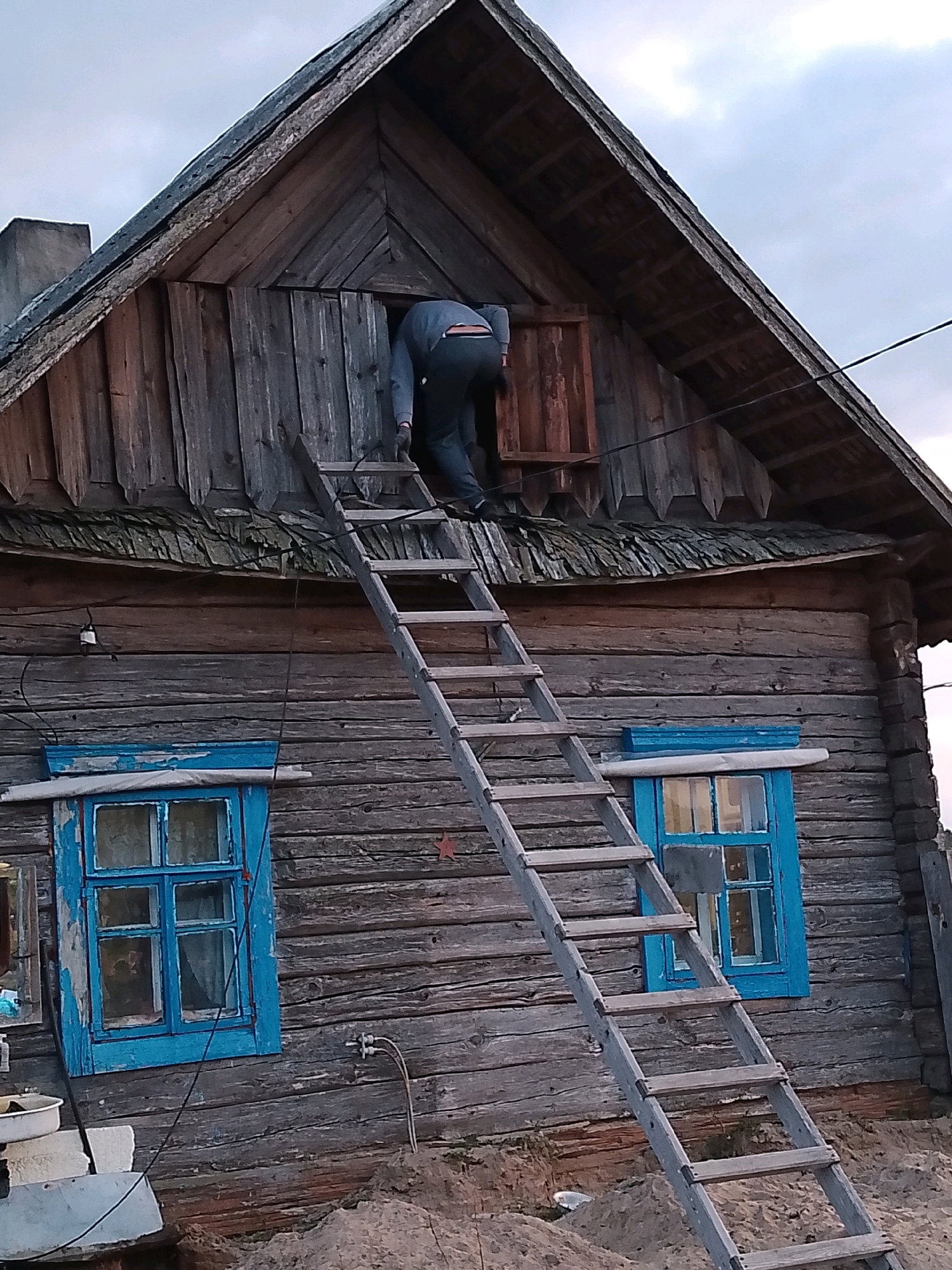 A person is climbing a ladder into the attic of a wooden house. The house is weathered and appears to be old. There are two windows with blue frames on the side of the house. The sky is overcast. The scene is likely rural, possibly in the countryside. The location is identified as Ленінскі сельскі Савет, Belarus.