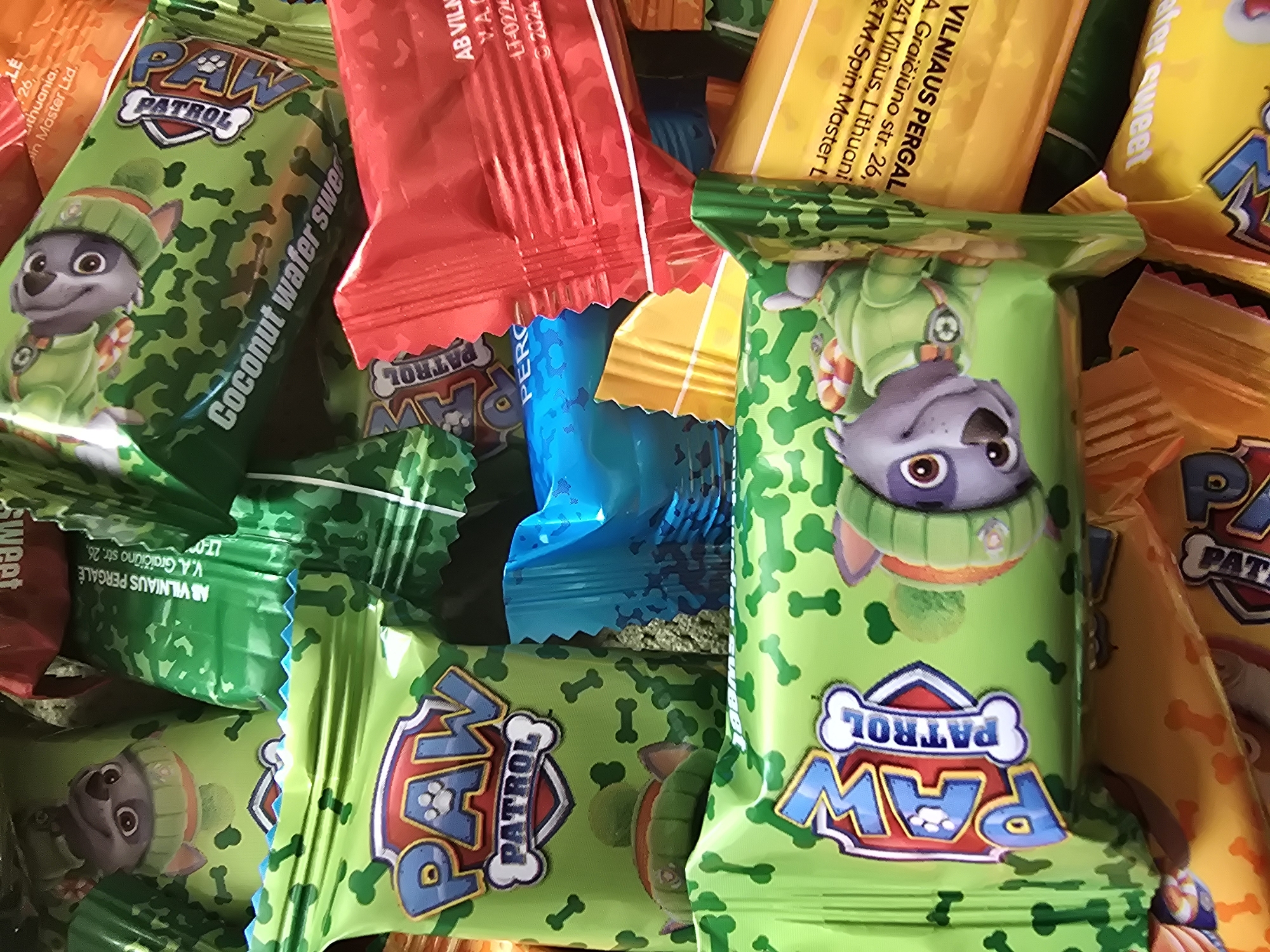 The image shows a pile of individually wrapped Paw Patrol coconut wafer sweets. The wrappers are predominantly green with a pattern of dog bones and feature characters from the animated series. Other wrappers in the pile are red, blue, yellow, and orange. Text on some wrappers includes "VILNIAUS PERGALE," "V.A. Graičiūno str. 26," "Lithuania," "Coconut wafer sweet," and "Spin Master Ltd." indicating the origin or manufacturer of the sweets. The scene is a close-up shot, highlighting the colorful packaging.