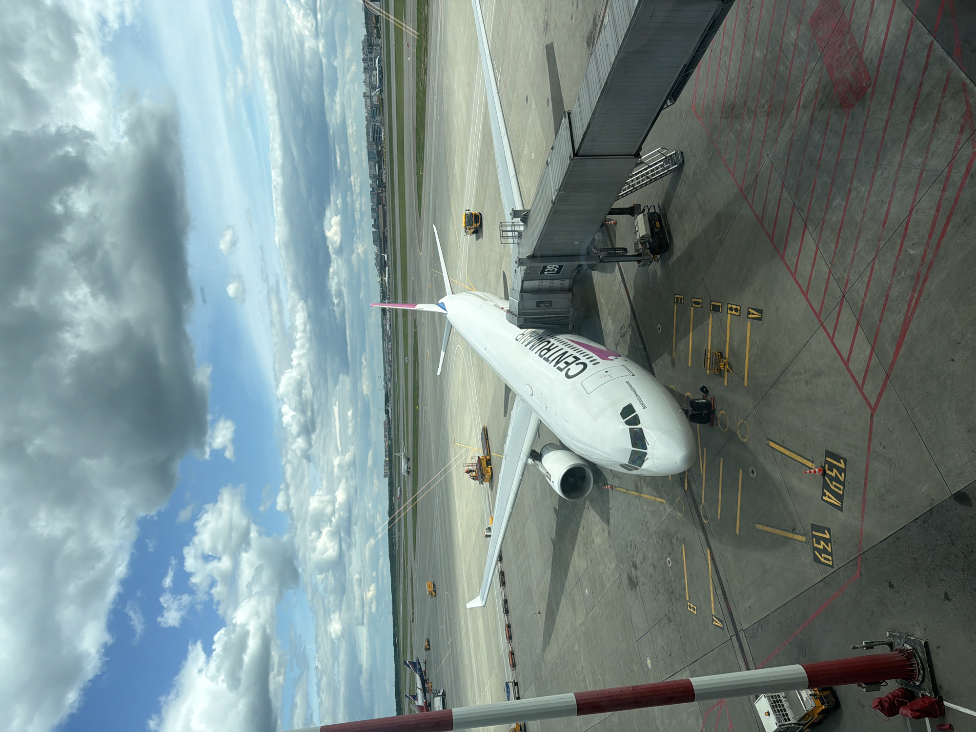 The image depicts an aerial view of an airplane, labeled "CENTRALNIY," connected to a boarding bridge at Sheremetyevo International Airport in Khimki, Russia. The airplane is white with a pink tail. The boarding bridge is grey and connected to a building. The ground is concrete with yellow and red markings indicating parking spots and taxiways. The sky is visible with white clouds and blue patches. The city of Khimki is visible in the background.