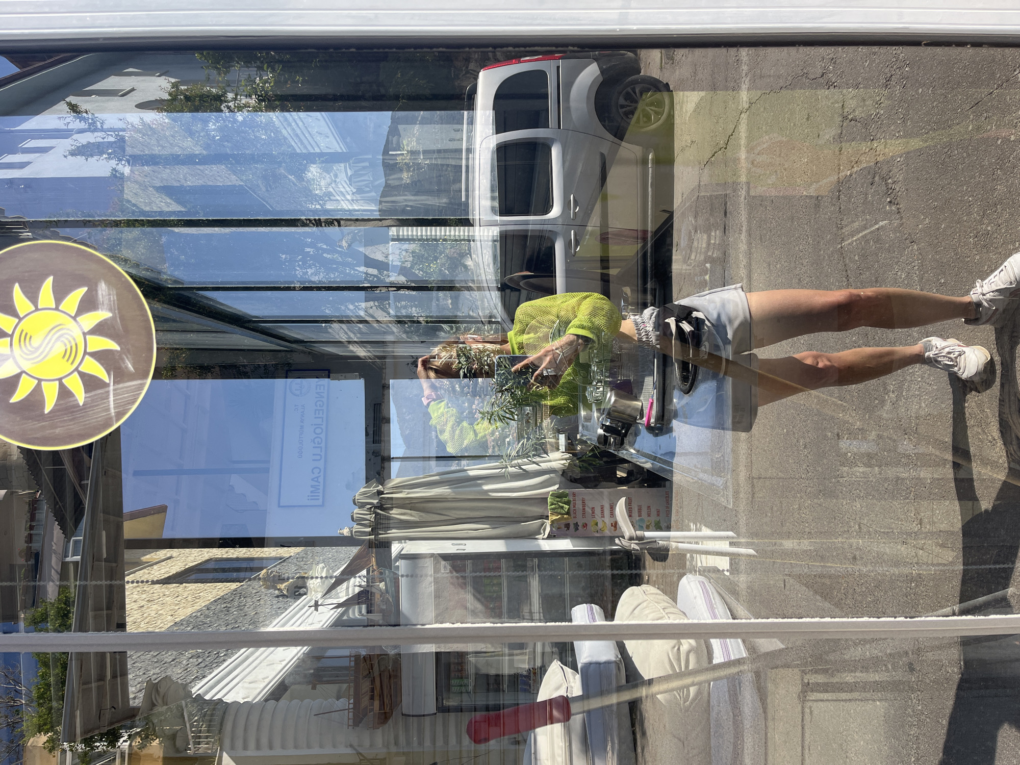 The image captures a reflection in a window, presenting a complex layering of scenes both inside and outside a cafe in Alanya, Turkey. The cafe's interior features cushions and a menu advertising flavors like "BLACK MULBERRY", "STRAWBERRY", "LEMON", and "BANANA", visible on a menu sign. A person, presumably the photographer, is reflected in the window taking a photo of their reflection. Outside, a white van is parked on the street, with elements of the surrounding buildings also reflected in the glass. A round sign bearing a yellow sun-like symbol is affixed to the window.