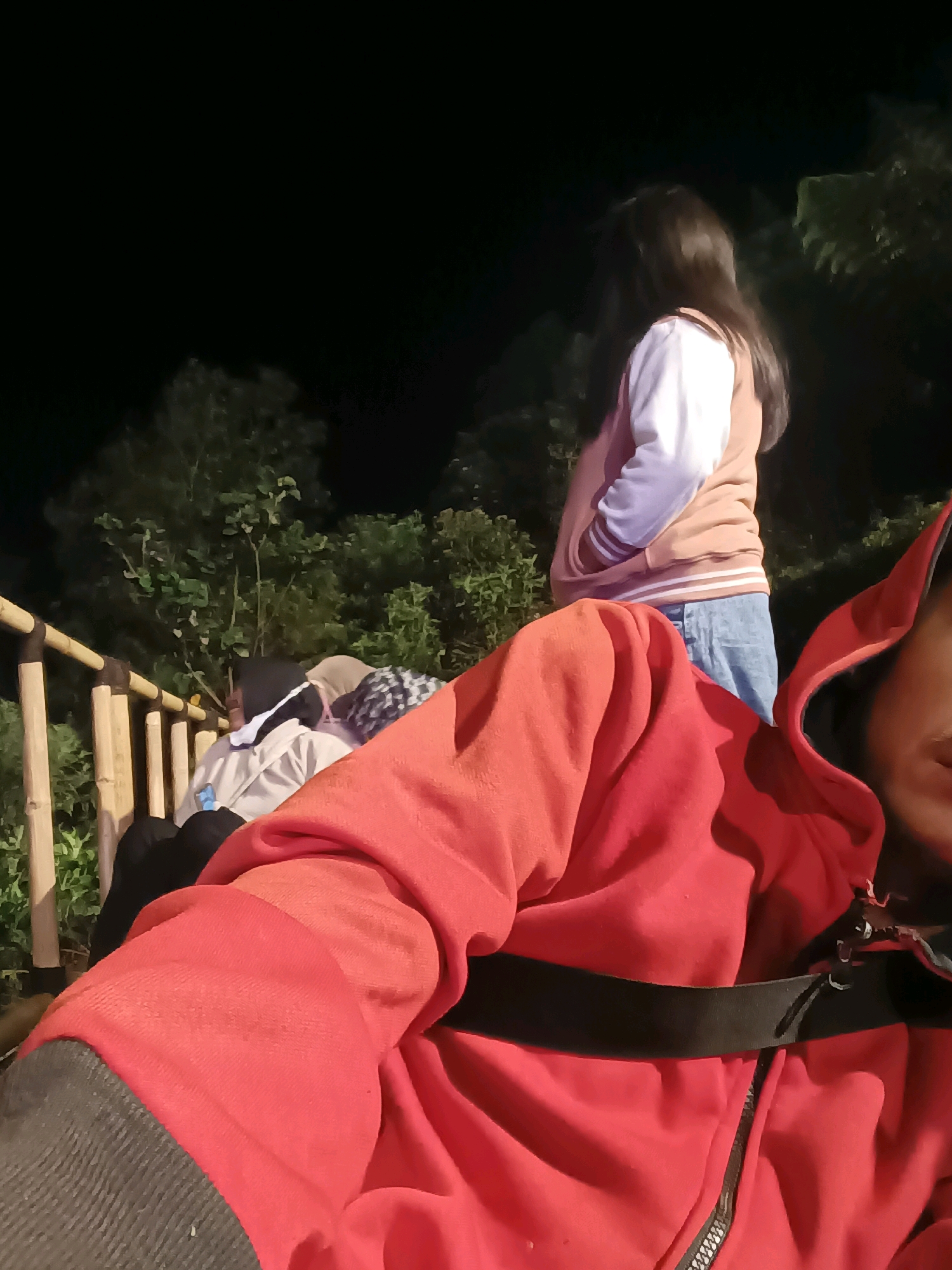 A close-up, low-light, first-person perspective shot shows a person in a red hooded jacket with a black strap across the chest. The camera is positioned at a slight downward angle, capturing the texture of the jacket and a partial view of the wearer's face. In the background, out of focus, a woman with long dark hair stands facing away from the camera, wearing a light pink and white bomber jacket and blue jeans. To the left, a bamboo railing separates the foreground from a group of people seated, some wearing headscarves. Green foliage and trees are visible behind them, and the overall setting appears to be outdoors at night. The location is identified as Keditan, Indonesia.