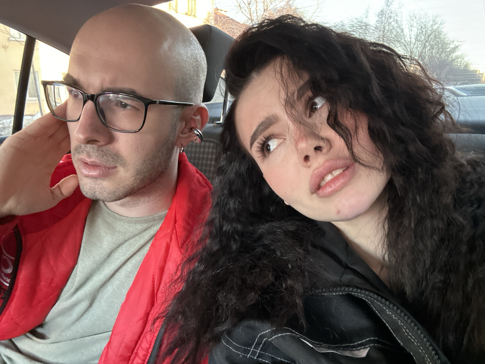 Two individuals are captured in what appears to be the interior of a car. The person on the left is a bald male wearing glasses, a grey t-shirt, and a red jacket. He has an earring in his left ear. His hand is raised to his left cheek. The person on the right is a female with long, dark, curly hair. She is wearing a dark jacket. She is looking up and to her left. The background shows trees without leaves and what looks like other cars. Outside the car window on the left is a yellow building. The location is Kazan, Russia.