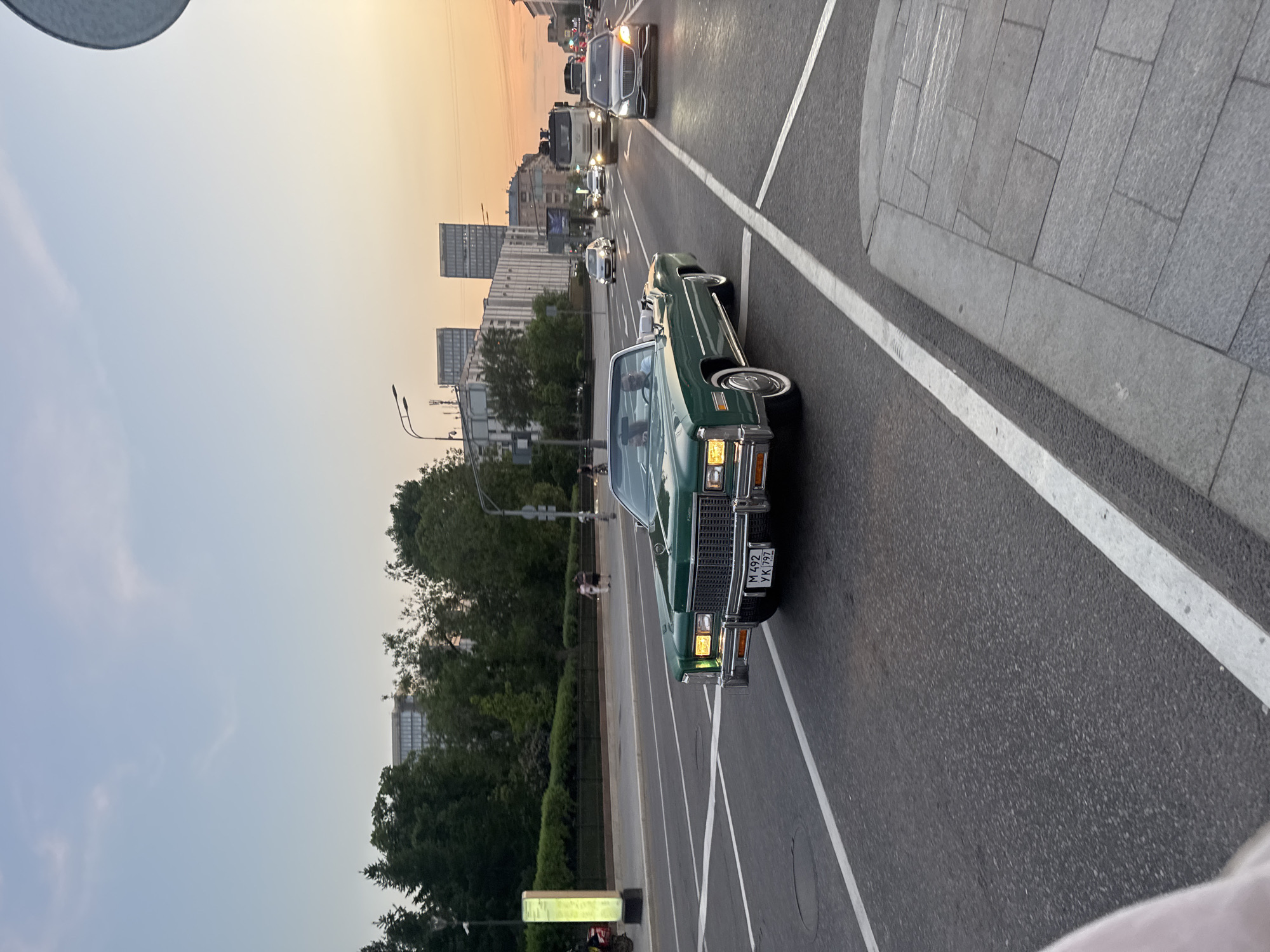 The image depicts a green vintage car driving on a multi-lane road in Moscow, Russia. Several other vehicles are visible in the background, including cars and what appears to be a truck. The road has white lane markings and a sidewalk is visible on the right. Buildings are present in the background, suggesting an urban setting. The sky is light, suggesting it may be near dusk or dawn. There are trees visible between the road and the buildings. A license plate is visible on the front of the car. People can be seen walking in the background.