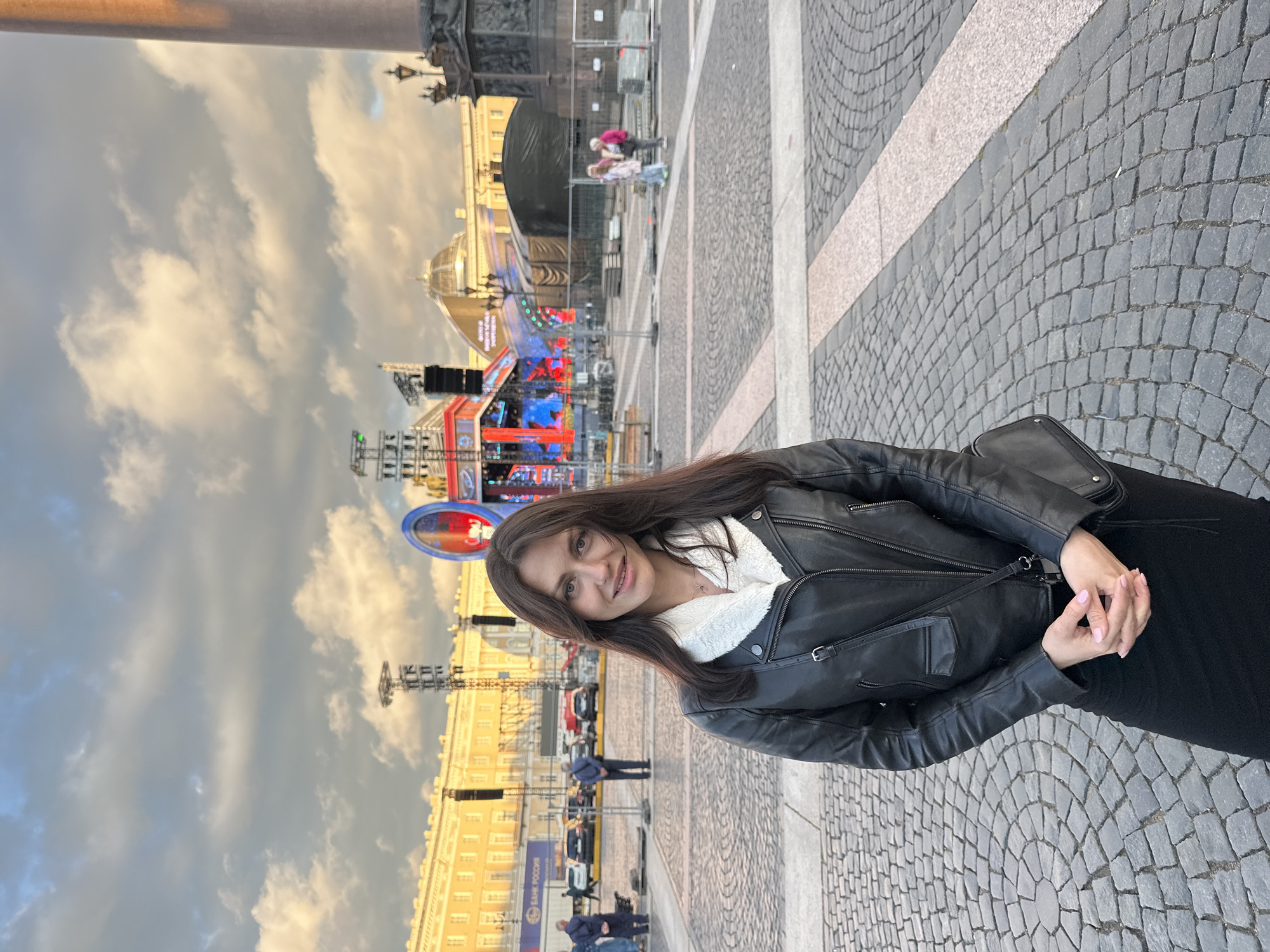 A young woman with long, dark hair stands in the foreground, looking towards the camera. She is wearing a black leather jacket with a white fur collar, and a black skirt. Her hands are clasped in front of her. Behind her, a large, open plaza is paved with cobblestones. In the background, a grand, yellow building with numerous windows is visible, along with a stage set up with lights and screens. A large column stands to the right of the frame. Several cars and a few people are scattered throughout the plaza. The sky above is cloudy. The location is likely Palace Square in Saint Petersburg, Russia, as indicated by the architectural style of the buildings and the presence of the Alexander Column.