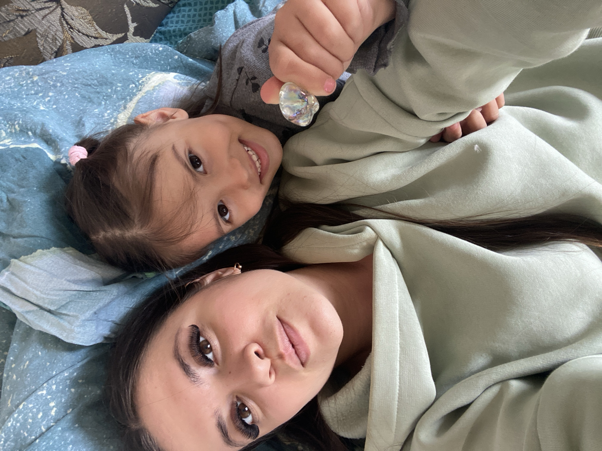 Two people, likely a mother and daughter, are lying close together on a patterned blue blanket. The daughter, wearing a grey top, is holding a clear, prism-like object in her hand. The mother is wearing a light green hoodie. They are indoors, likely in a home setting. No evidence supports the location being Zhigalki, Russia.