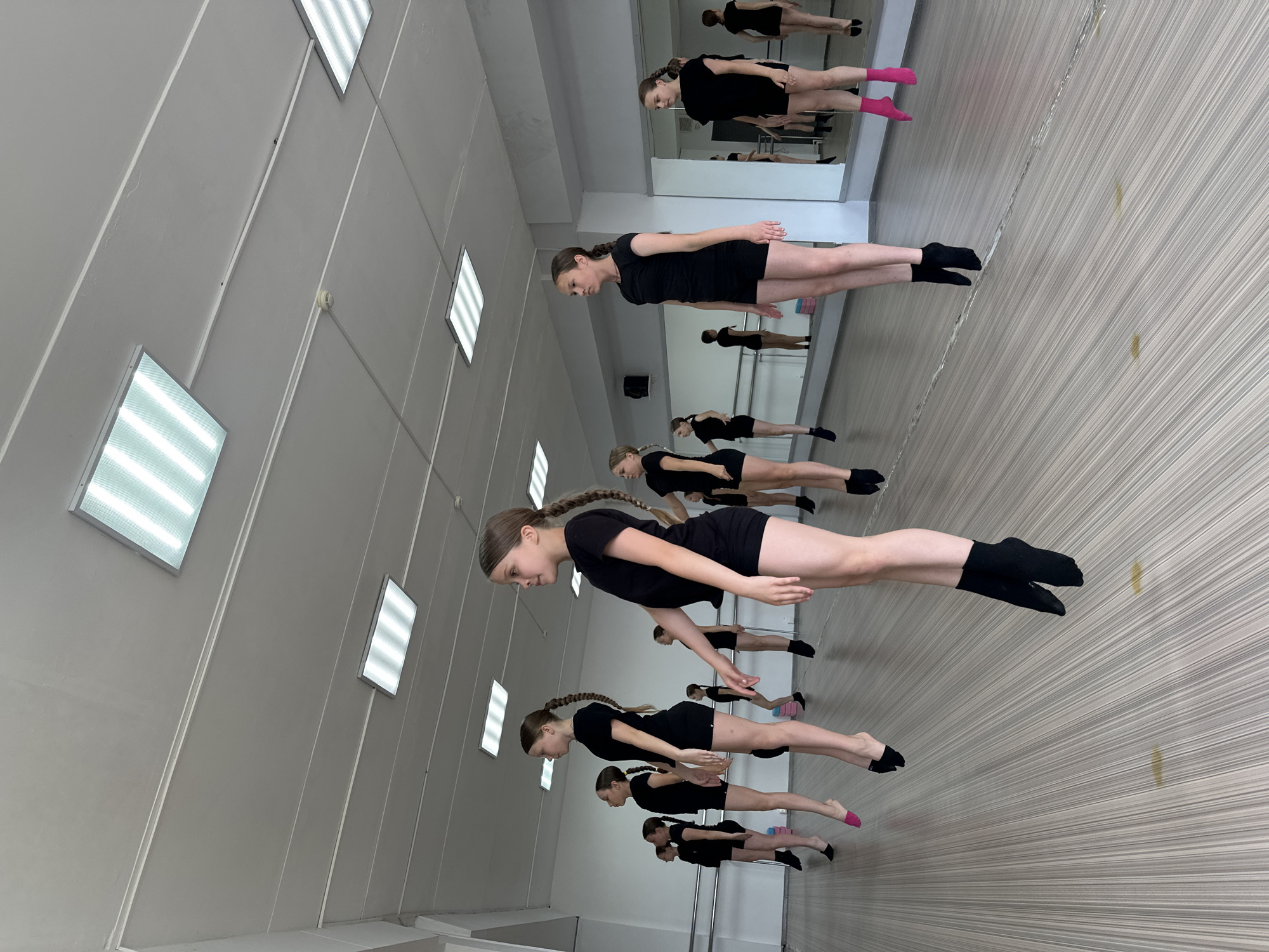 A group of young girls in black leotards and shorts are practicing ballet in a studio. They are all in mid-air, appearing to be jumping or performing a levitation pose. The studio has a wooden floor, mirrors along one wall, and fluorescent lights on the ceiling. The girls have their hair in braids and are wearing ballet socks. The scene is well-lit and appears to be a professional dance studio. This image was taken in a dance studio in Votkinsk, Russia.