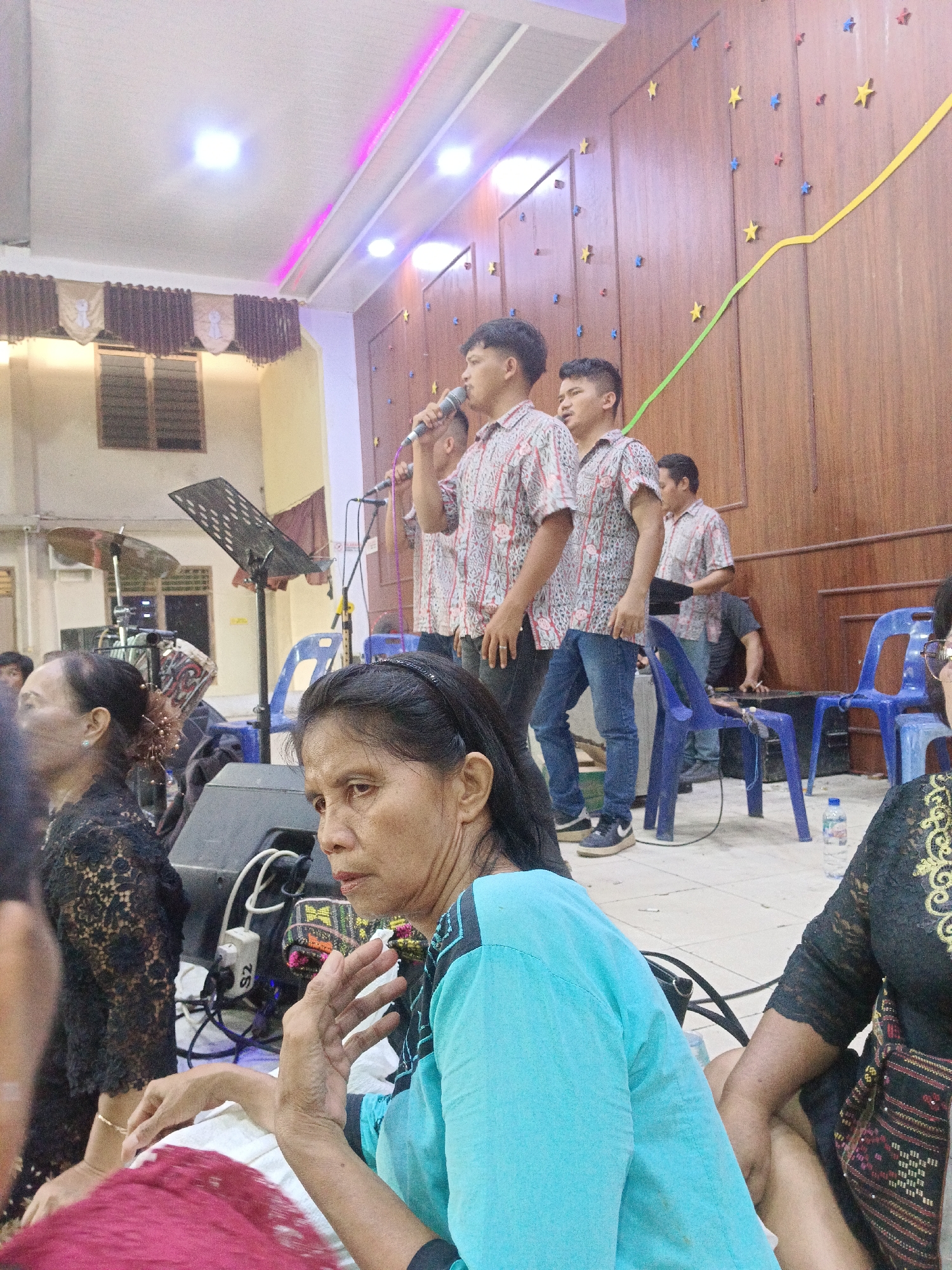The image depicts an indoor gathering, likely a church service or event, in Aek Kanopan, Indonesia. In the foreground, two women are visible, one in a bright turquoise top with dark accents and the other in a black lace top. The woman in turquoise is in focus, with her hand raised as if gesturing or speaking. Behind them, three young men in patterned shirts and jeans are standing on a raised platform, with one holding a microphone. They appear to be singing or performing. The background features a wooden paneled wall adorned with colorful star cutouts and a yellow ribbon, suggesting a festive or celebratory atmosphere. There are also several blue plastic chairs, musical equipment including a drum set and a music stand, and a bottle of water on the floor. The lighting suggests it is daytime, with artificial lights and natural light from windows in the background. The overall scene conveys a sense of community and shared experience.