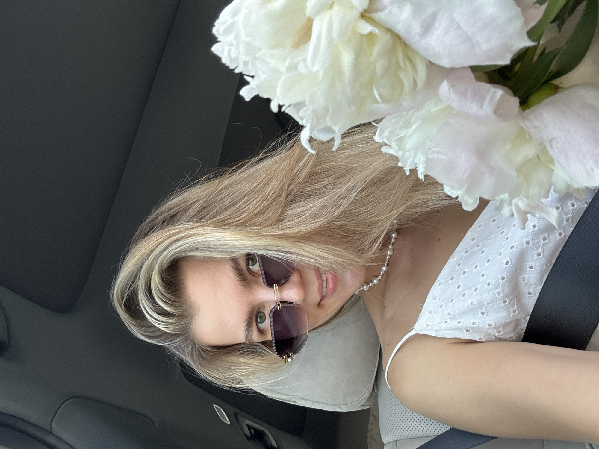 A young woman with blonde hair and green eyes smiles at the camera, wearing sunglasses and a pearl necklace. She is dressed in a white eyelet camisole and is holding a bouquet of white peonies. The woman is sitting in a car, as evidenced by the headrest and seatbelt visible in the frame. The background is dark, suggesting the interior of the car, with a hint of the outside world possibly visible through a window. The lighting is soft and natural, indicating it is daytime. The scene is captured from a slightly low angle, looking up at the woman. The overall impression is one of a casual, sunny day outing.