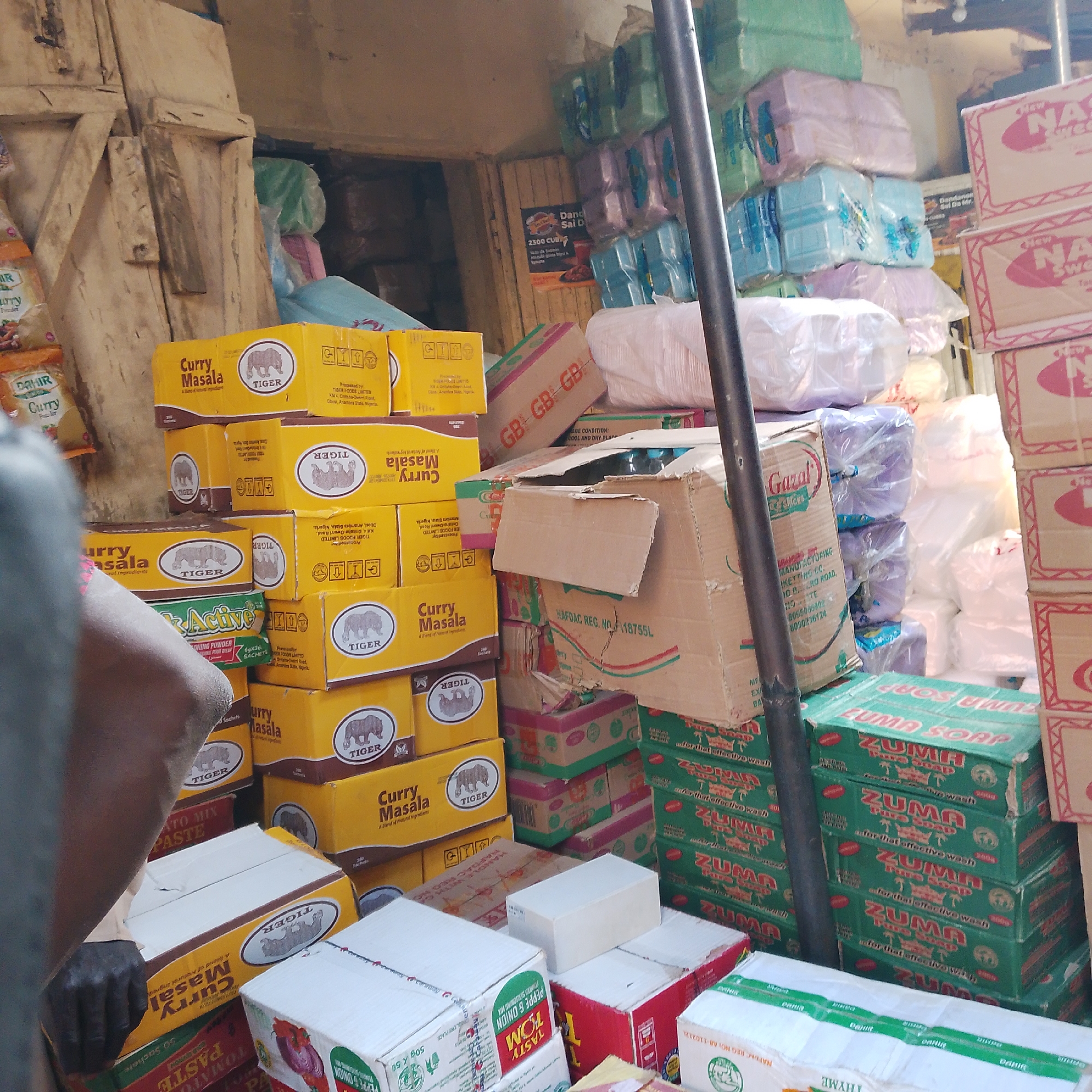 The image shows a retail stall, likely a market, in Arawa, Nigeria, with a focus on stacked goods. In the foreground, a person's arm and shoulder are visible on the left, partially obscuring some of the products. The stall is filled with various boxed items, predominantly food products like "Curry Masala" and "Tomato Mix Paste" from brands like Tiger and Dahir, displayed in yellow and red packaging. There are also stacks of green boxes labeled "ZUMA SOAP". Other items include stacks of what appear to be toiletries or household goods wrapped in plastic, in various pastel colors, and some larger cardboard boxes, one of which is open. The background is dimly lit, suggesting an indoor or covered space. The overall impression is of a well-stocked, busy market stall.