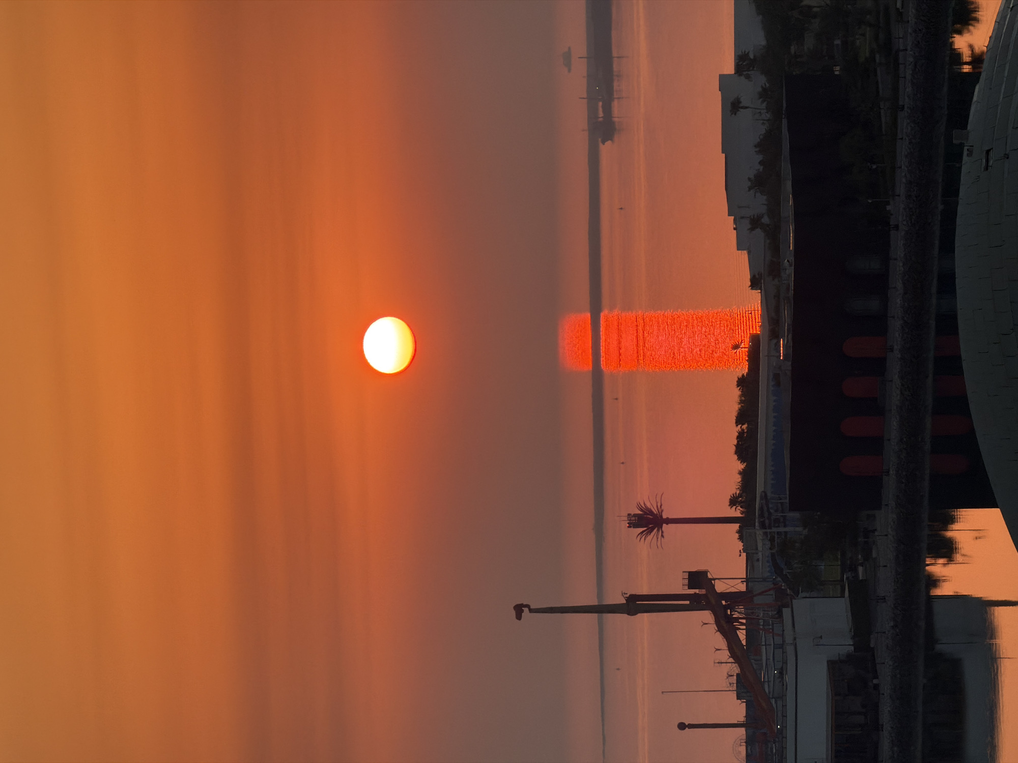 The image depicts a vibrant sunset over a body of water, casting an intense orange glow across the sky and reflecting vibrantly on the water's surface. A bright, full sun is positioned in the upper center of the frame. In the foreground, there are various structures, including buildings and industrial elements, as well as a body of water. A tall, thin structure with a light at the top is visible on the left, along with a palm tree silhouette on the right. Distant ships and a hazy shoreline are visible on the horizon. The overall scene suggests an urban or industrial waterfront setting at dusk.