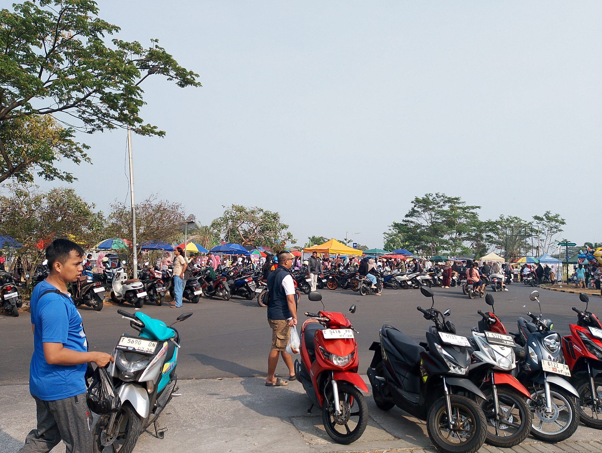 The image depicts an outdoor market scene in Karawang, Indonesia, with numerous parked motorcycles forming a significant part of the foreground and midground. The market stalls, indicated by colorful canopies, are visible in the background, suggesting a commercial activity. Several individuals are present, some walking and others appearing to be browsing or interacting. A man in a blue t-shirt and grey pants is walking in the left foreground, holding a black plastic bag. Another man in a white t-shirt and khaki shorts is walking in the middle ground. The motorcycles are predominantly scooters, with varying colors and models, suggesting a common mode of transportation in the area. The environment appears to be a paved area, possibly a parking lot or open space adjacent to the market. Trees and vegetation are present in the background and on the left side of the image, indicating an outdoor setting. The sky is overcast, suggesting a daytime scene.