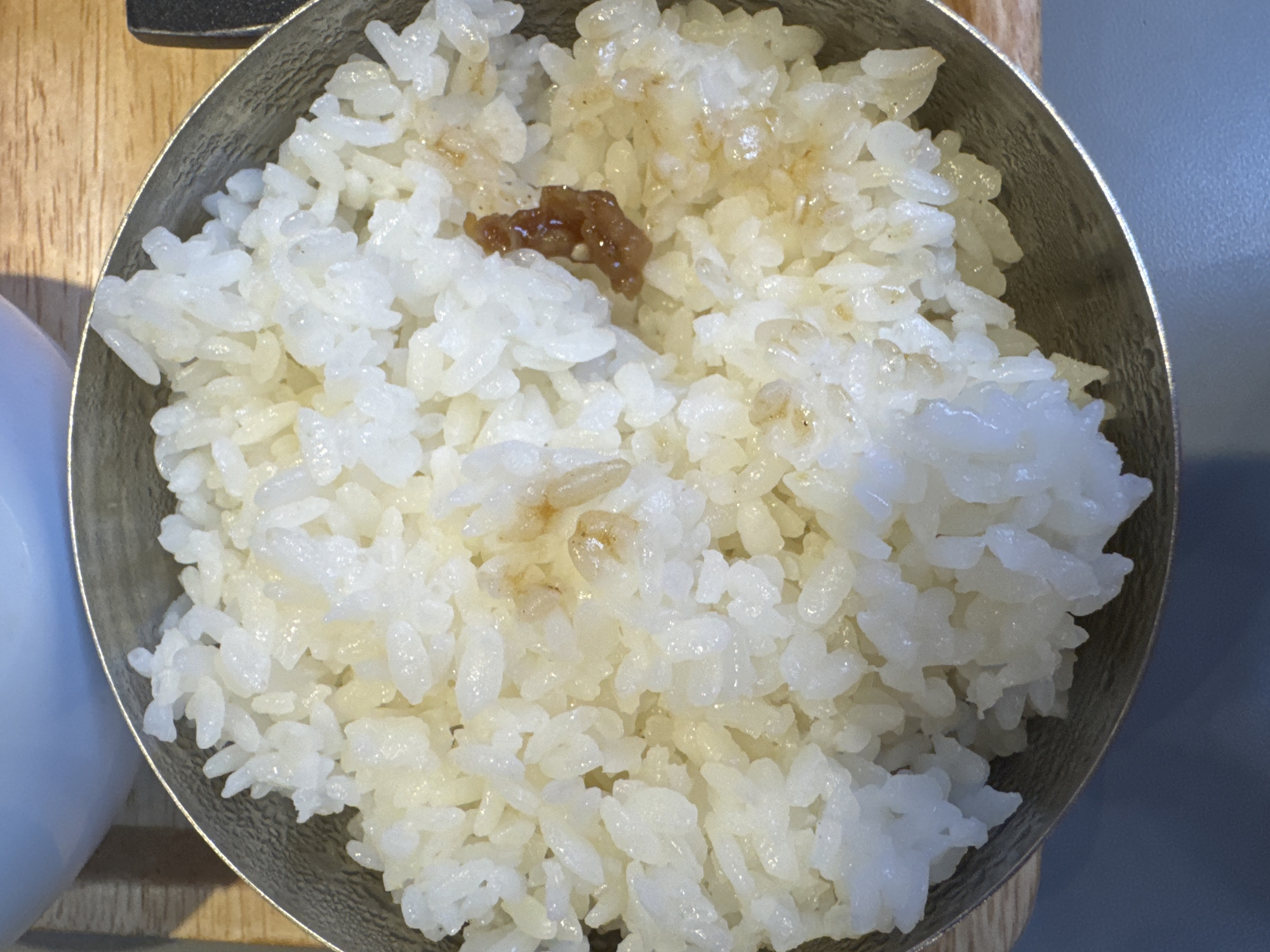 A close-up, overhead shot captures a portion of a metallic bowl filled with steamed white rice. A small, dark brown piece of food, possibly a piece of meat or a seasoning, rests on top of the rice. The rice appears slightly glossy, indicating it is freshly cooked. The background shows a wooden surface and a hint of a light-colored bowl or dish. The image is a food photography shot, likely taken in a restaurant. There is no indication of the city of Bangkok, Thailand, in the image.