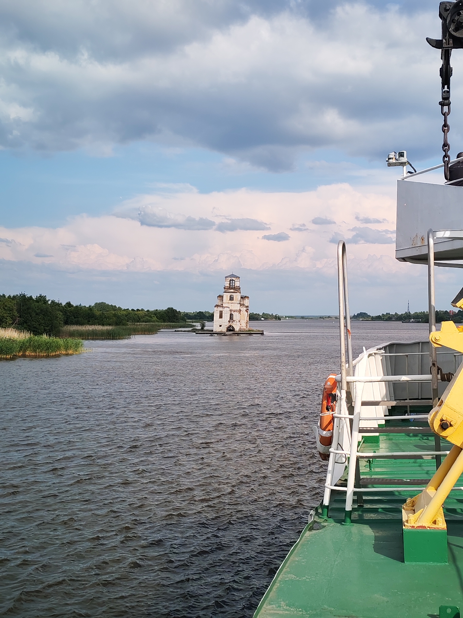 The image shows a body of water with a white, multi-story structure on a small island in the distance, identified as the Church of the Nativity of the Virgin Mary in Krokhino, Russia. The church appears weathered and partially ruined. In the foreground, the green deck and railing of a vessel are visible, along with a bright yellow mechanical arm and an orange life preserver. The sky is partly cloudy, with a mix of blue and gray tones. The water is choppy, indicating a breezy day. The overall scene suggests a view from a boat traveling on a river or lake towards the historical landmark.