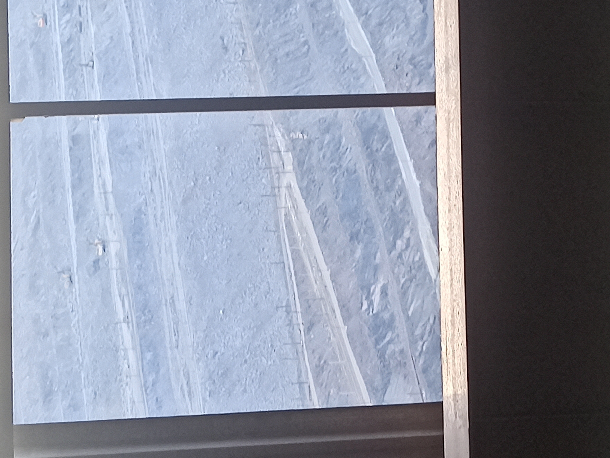 The image shows an aerial or elevated view of what appears to be a construction site or a large industrial area. The ground is covered in a light blue or white material, possibly snow, sand, or a protective tarp. There are several parallel, raised pathways or structures running across the scene. Small, dark, vertical objects are spaced out along these pathways, resembling poles or markers. In the distance, there are rough, uneven textures that suggest piles of material or hilly terrain.
In the upper left quadrant, there are faint, light-colored shapes that could be small structures or vehicles. Further down the pathways, more of these small, ambiguous shapes can be seen, some appearing to be people or machinery. The overall impression is one of a large-scale operation taking place outdoors.
The light in the image is diffused, suggesting it could be daytime, but the weather conditions are unclear. The colors are predominantly cool blues and whites, with some darker areas on the right side of the image, which appear to be a shadow or the edge of a structure. The perspective is looking down and across the scene, with a wooden frame or window sill visible on the right side. There is no visible text in the image.