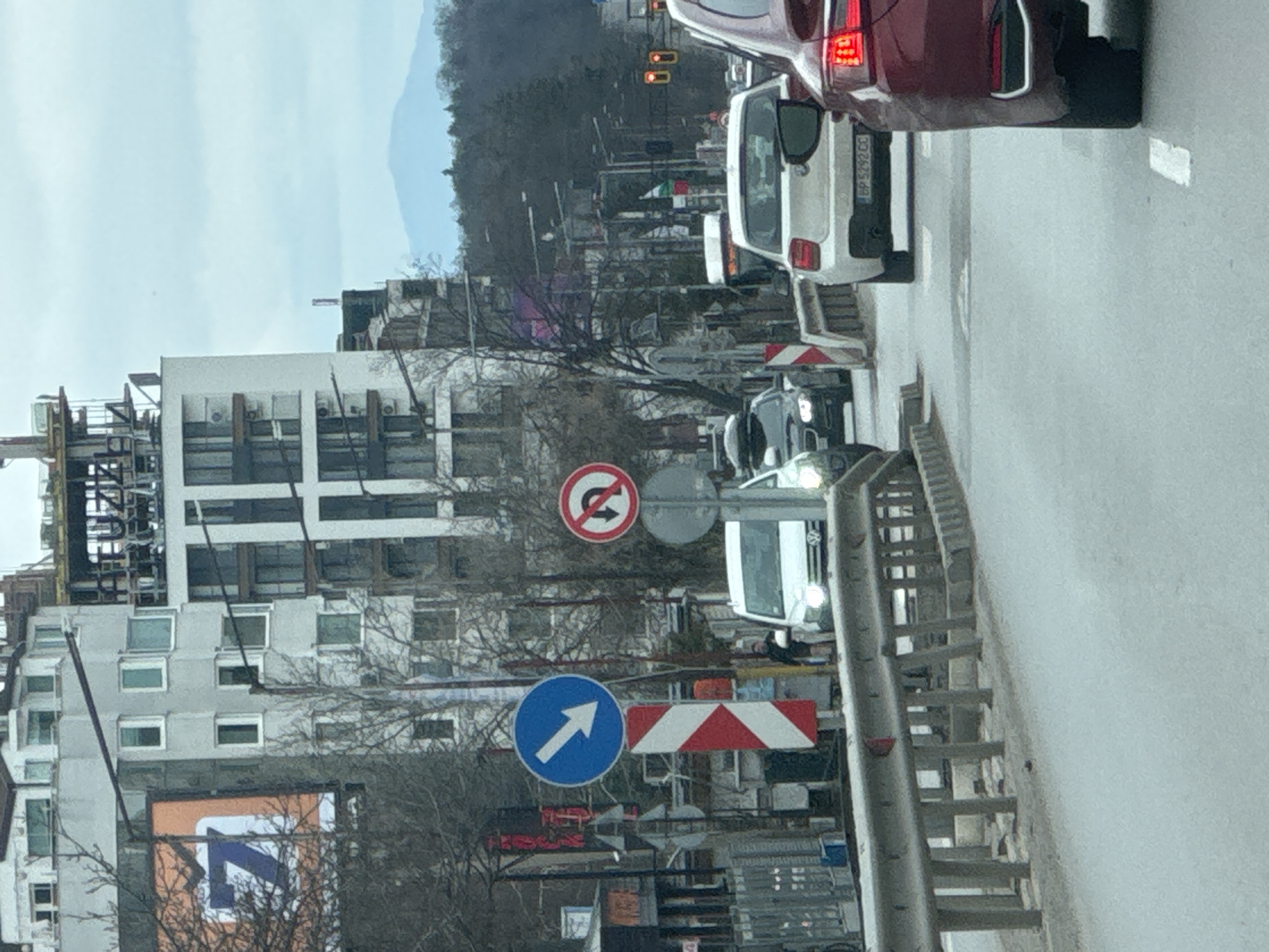 The image captures a street scene in Sofia, Bulgaria. In the foreground is a gray metal guardrail, partially obscuring road construction. Traffic signs indicating "no U-turn" and "turn right" are visible among orange and white construction barriers. A line of cars is stopped at a traffic light, including a maroon SUV in the top right and a white van in the middle. In the background, a tall building, possibly under construction or renovation, dominates the left side of the frame. Bare trees fill the space between the building and the traffic. Further back, a distant mountain range is visible under a cloudy sky.
