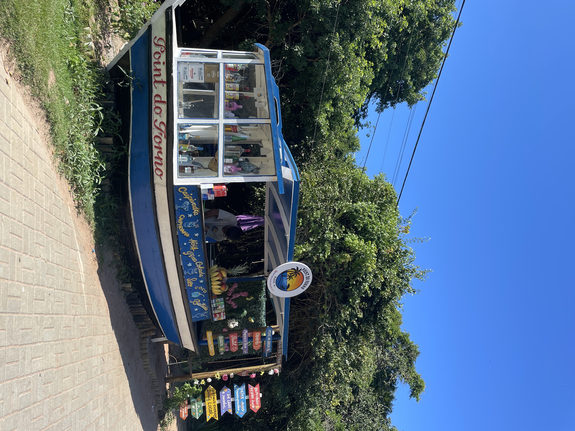 The image displays a brightly lit outdoor scene featuring a boat-shaped kiosk under a clear blue sky, likely in Armação dos Búzios, Brazil, specifically near Forno Beach. The kiosk, painted white and blue, serves as a vendor stand for drinks and snacks.
The main subject is this unique kiosk, positioned on a paved pathway bordered by green foliage and trees. The front part of the kiosk, appearing to be the bow of a boat, has "Point do Forno" written in red script. Behind this, a glass display case showcases various bottles of spirits and other items. A person is partially visible inside the kiosk, suggesting active business. Below the display, bottles of Red Bull are visible. Fresh fruits, including bananas and pineapple, are displayed on the counter.
The kiosk offers a variety of beverages and food, as indicated by hand-painted signs on its side, including "Caipivodka," "Caipirinha," "Água" (Water), "Pina Colada," "Sorvete" (Ice Cream), "Coco" (Coconut water), and "Batida" (a type of Brazilian cocktail/shake).
A prominent circular sign attached to the kiosk features a sunset palm tree logo and the text "Point do Forno BUZIOS." Below this, a cluster of colorful, arrow-shaped wooden signs points in various directions, listing food items and motivational phrases. Food items include "PRAIA DO FORNO" (Forno Beach), "PEIXE NA BRASA" (Grilled Fish), "Camarão" (Shrimp), "Moqueca & Peixe," "Pastel" (Pastry), "BATATA" (Potato), and "Churrasco Misto" (Mixed Grill). A longer sign reads: "BEM-VINDO RESPIRE fundo DEIXE suas Preocupacões DE LADO e receba as boas ENERGIAS Deste LUGAR" (Welcome, breathe deep, let go of your worries and receive the good energies of this place). Another small sign says "pointdofornobeach."
The setting is outdoors on a sunny day, indicated by the bright lighting, sharp shadows, and clear blue sky with no clouds. Power lines are visible against the sky in the upper right. The ground is a light-colored paved surface, and lush green trees form a dense backdrop, suggesting a natural, possibly beachside, environment. The overall impression is that of a cheerful, tropical, and inviting spot for refreshments.
