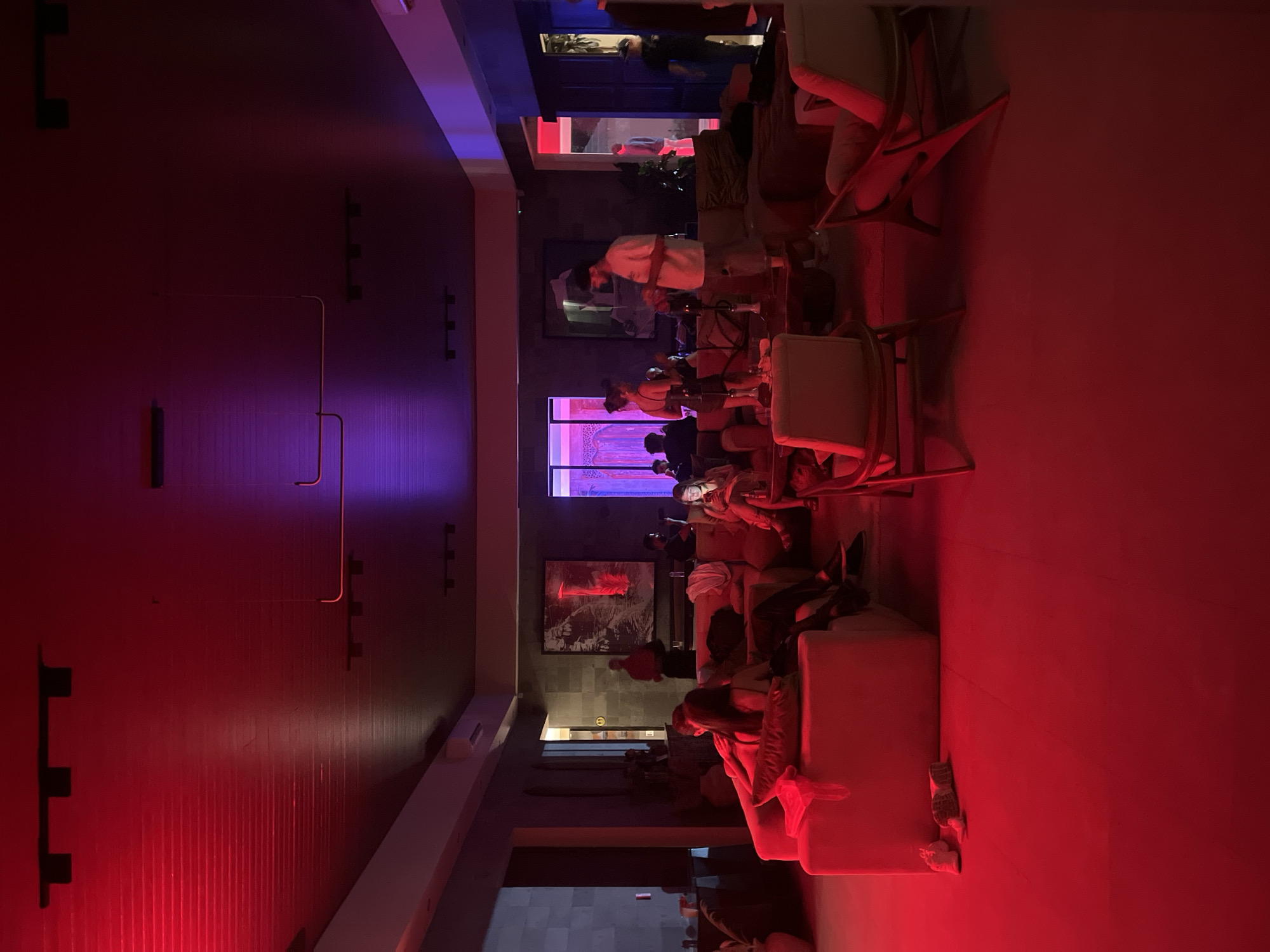 This image depicts an indoor lounge or bar area located in Kuta Selatan, Indonesia. The environment is characterized by atmospheric red and purple lighting. The entire floor and the lower half of the room are saturated with deep red light, which transitions to purple on the upper walls and ceiling, particularly towards the right side.
Approximately 15-20 individuals are present in the scene. In the left foreground, a couple is seated closely on a light-colored sofa. Next to them, another person is looking down at a smartphone, its screen illuminating their face. In the mid-ground, a man wearing a white t-shirt stands near a low table with a hookah, appearing to handle the device, with a woman standing close by. Other patrons are distributed across various sofas and chairs throughout the space, engaged in conversation or relaxation. A person in a red shirt, possibly staff, stands in the background near a large framed artwork on the rear wall. Several more individuals are visible standing and moving near open doorways on the right side of the frame.
The furniture includes multiple sofas of varying colors and several wooden-framed armchairs, particularly prominent in the foreground. Two pairs of casual athletic shoes are placed on the red-lit floor in front of a sofa on the left. Small tables are interspersed among the seating arrangements, some holding hookahs. The walls are dark and textured, adorned with large framed artistic prints. The ceiling is dark and features linear track lighting fixtures.