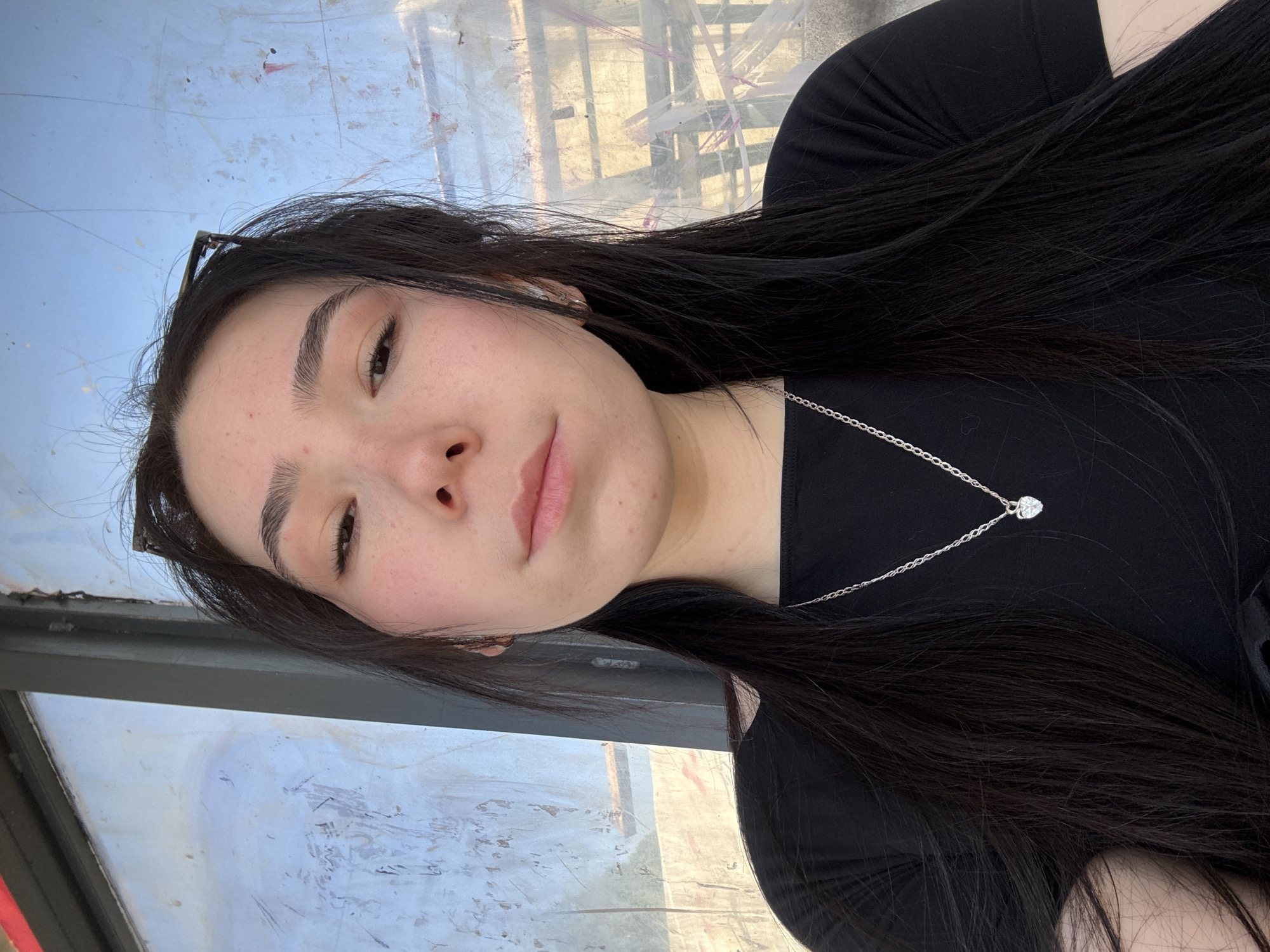 A young woman with long, dark hair and light skin is looking at the camera. She is wearing a black shirt and a silver necklace with a heart-shaped pendant. The background appears to be a bus stop with a grimy window in the city of Izhevsk, Russia. There is a pair of sunglasses propped on her head. The woman is facing sideways and has a light, pink lipstick.