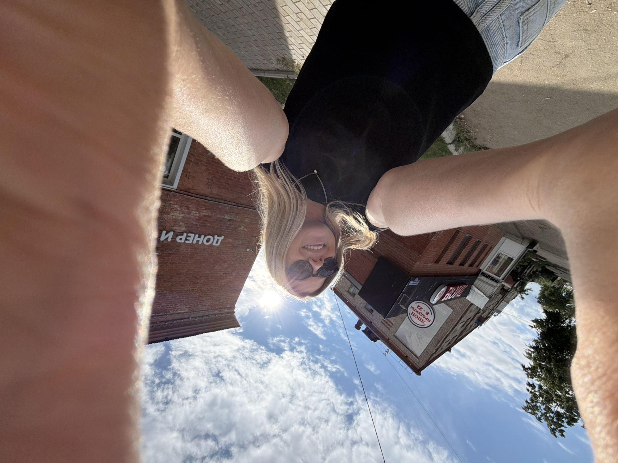 The image is a wide-angle, low-angle selfie taken outdoors in the city of Chaikovsky, Russia, on a sunny day with a bright blue sky and scattered white clouds. The foreground is dominated by the arms and shoulder of the person taking the photo, with a portion of their face visible. The person has blonde hair and is wearing sunglasses and a black top.
In the background, there are two brick buildings. The building on the left appears to be older, with a sign that reads "ЛЮЦИН ПРОДУКТЫ 8-22" (Lyutsyn Products 8-22) and another smaller sign. There are windows and doors visible on this building. The building on the right has a sign that reads "ДОНЕР И" (Doner I). The ground in front of the buildings is paved with light-colored bricks or tiles, and there is some greenery visible. The overall impression is of a street scene in a residential or commercial area.
