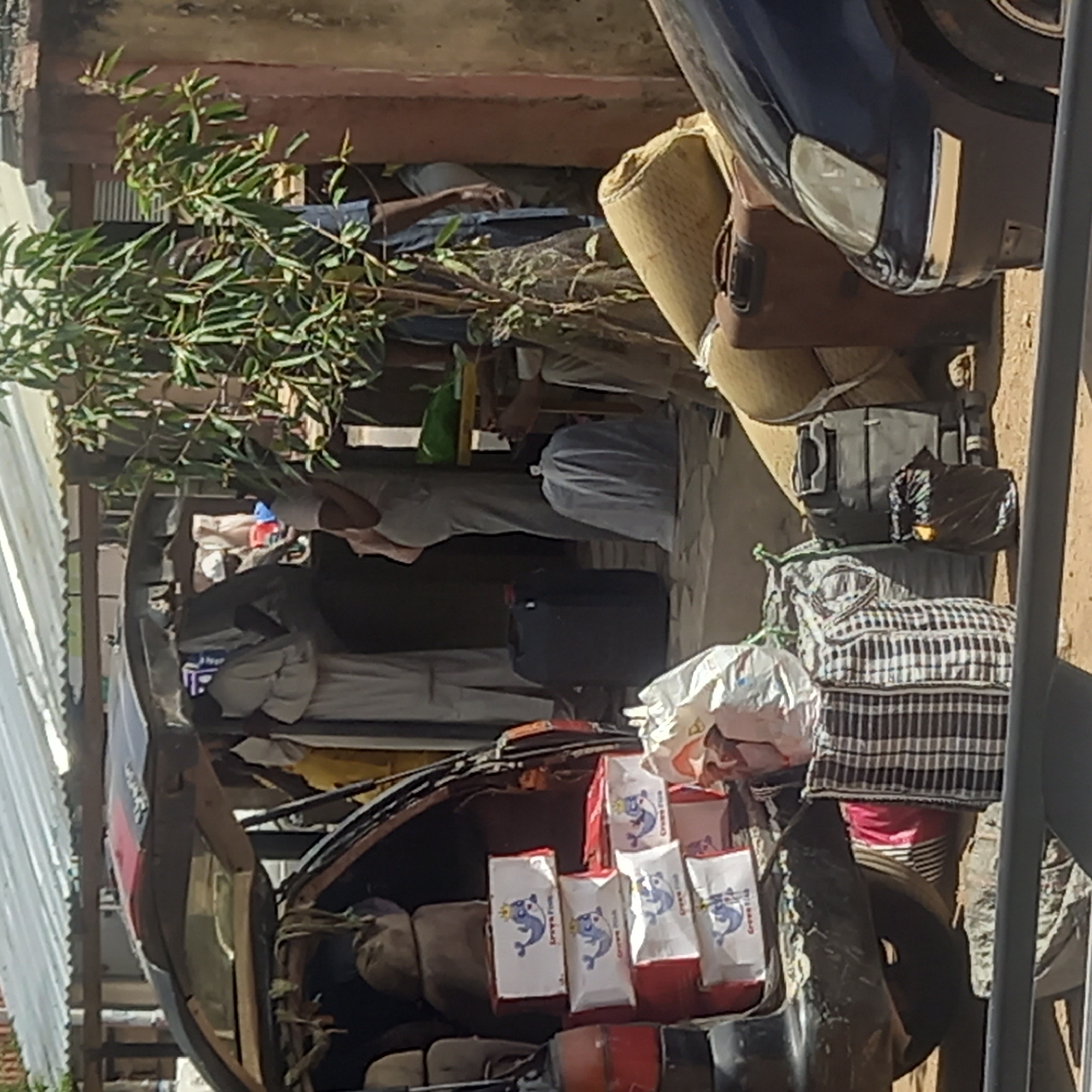 In Dadi Riba, Nigeria, the image shows a crowded outdoor scene. A damaged black and red vehicle is packed with various items, including bags with a blue dolphin logo. To the side, another vehicle, primarily blue, is also filled with items. In the background, a person in light-colored clothing stands near additional belongings. The surroundings include plants, corrugated metal sheets, and other structures.