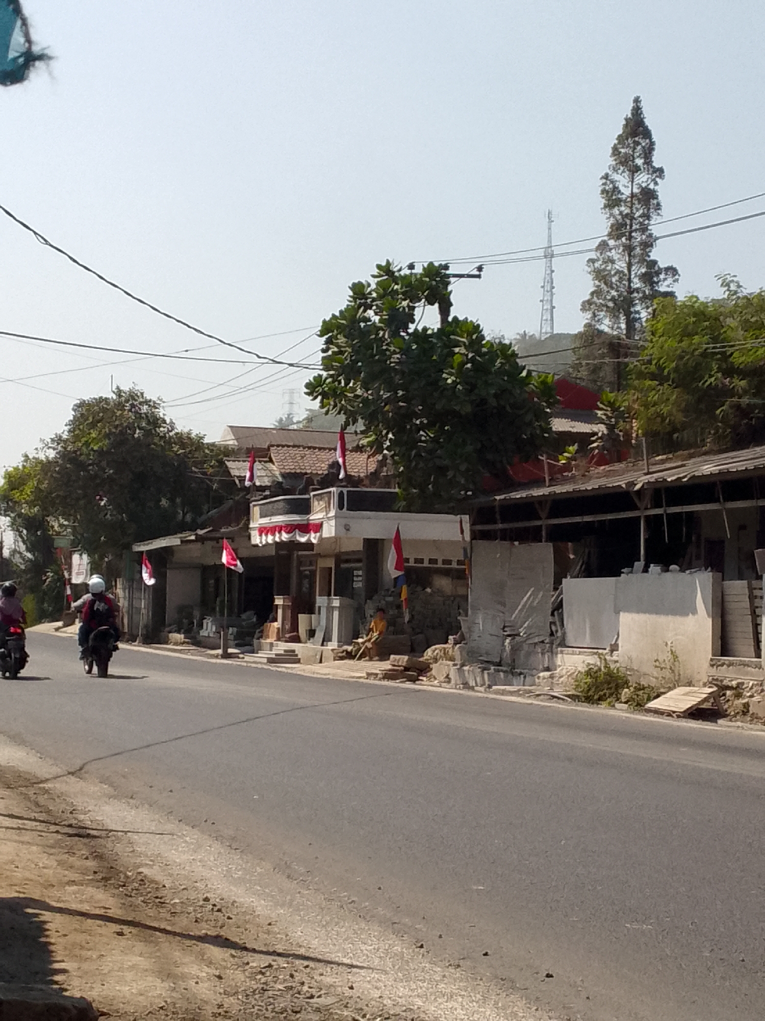 The image depicts a street scene in Gunungmasigit, Indonesia, under a bright, hazy sky. The foreground is dominated by a paved road with a gravelly shoulder. On the right side of the road, a row of low-rise buildings with corrugated metal roofs is visible. Some of these buildings appear to be shops or residences, with their facades decorated with red and white Indonesian flags, suggesting a national celebration or commemoration. In front of one building, stacked stone slabs and bricks are arranged, possibly for sale or construction. A person in a yellow shirt is seated near the stacked materials.
Further down the road, two motorcycles are traveling away from the camera. The motorcycle on the left has a rider and a passenger, both wearing helmets. The motorcycle on the right has a single rider, also wearing a helmet. Trees, some with dense foliage, line the area behind the buildings, and a tall, slender coniferous tree stands out on a distant hill, partially obscured by other greenery. A telecommunications tower is also visible in the background, adding to the urban or semi-urban landscape. Electrical wires crisscross the upper portion of the image. The overall impression is of a sunlit day in a developing area, with everyday activities taking place along the roadside.