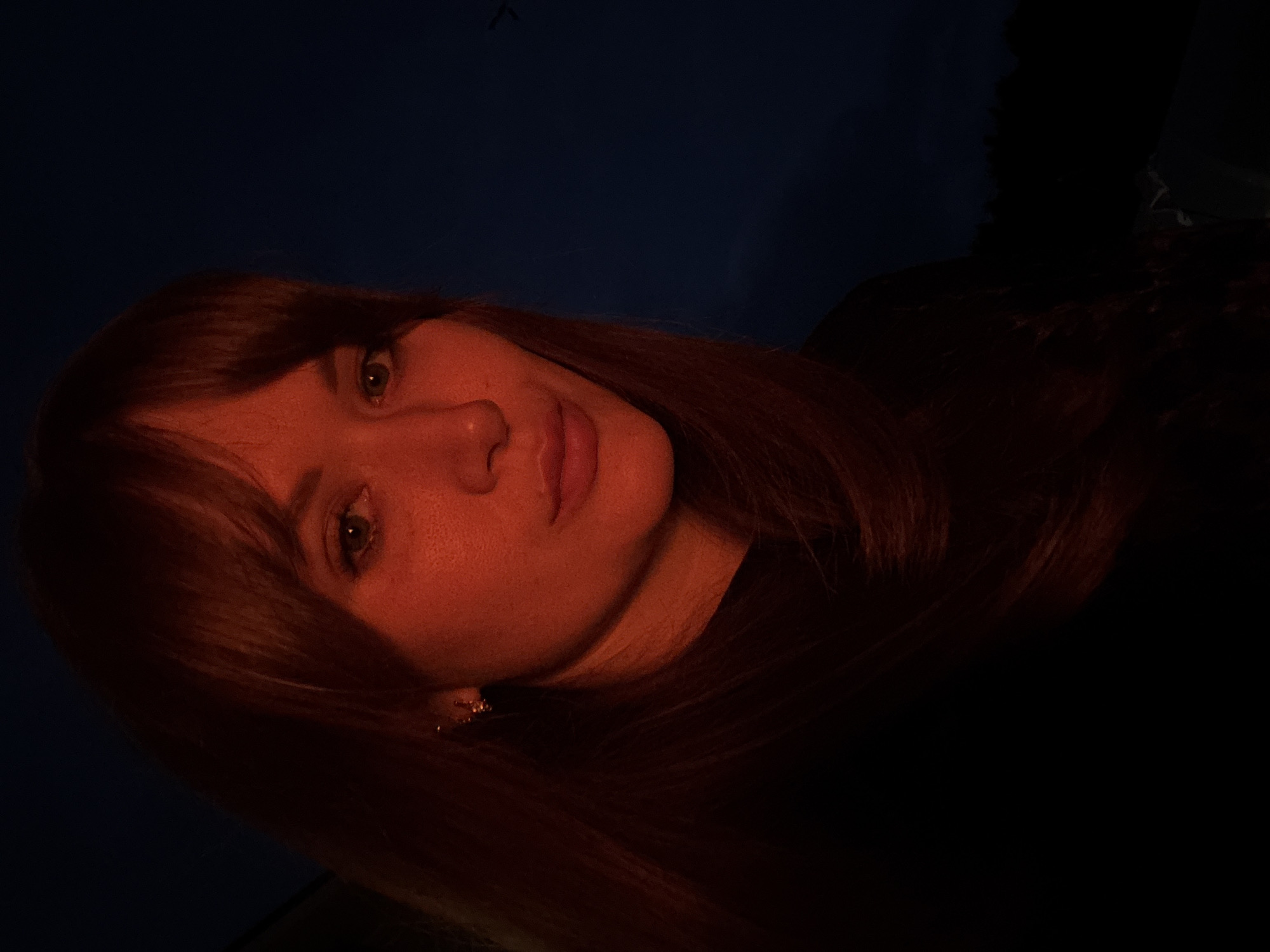 A close-up, self-portrait photograph shows a woman with auburn hair and bangs against a dark blue twilight sky. Her face is illuminated by warm, reddish-orange light, suggesting a nearby light source such as a fire or lamp. She is wearing a black shirt and a small hoop earring in her left ear. The background is mostly dark, with a faint silhouette of trees visible in the lower right corner. The image appears to be taken outdoors, possibly in the city of Chaikovsky, Russia, given the dark, natural environment visible in the background, which could be consistent with a rural or semi-rural setting within or on the outskirts of the city. There are no other people or objects of significance in the frame. The overall mood is intimate and slightly mysterious due to the dramatic lighting.
