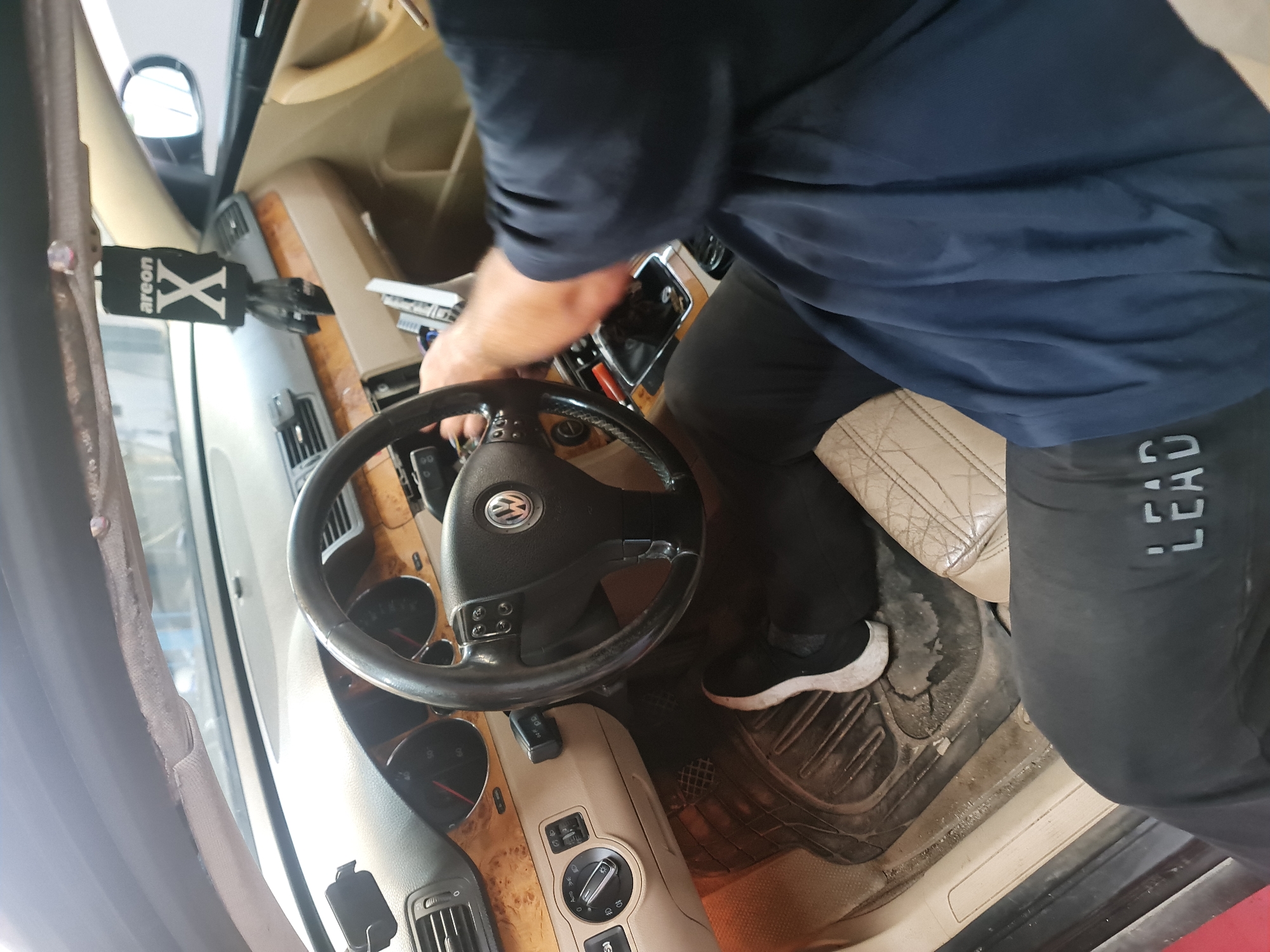 The image displays the interior of a car, specifically the driver's side, where a person is engaged in an activity. The perspective is from above and to the right, looking down into the vehicle.
A person is partially visible, wearing a dark blue t-shirt and black pants with the word "LEAD" printed vertically along the outer side of their left leg. They are wearing black and white sneakers. Their right arm extends towards the car's center console, where wires and what appears to be part of a car audio system or electronic component are exposed, suggesting maintenance, repair, or installation work is underway.
The car's interior features a black steering wheel with a Volkswagen (VW) logo at its center. The dashboard is adorned with a prominent wooden trim, housing air vents and gauges. A beige, quilted-pattern driver's seat shows signs of use. A black rubber floor mat below the person's feet appears dirty. Clipped to an air vent on the left side of the dashboard is a black car air freshener with "areon X" written on it. Natural light fills the cabin, indicating it is daytime. The setting suggests a workspace for automotive repair or customization, located in Buzovgrad, Bulgaria.