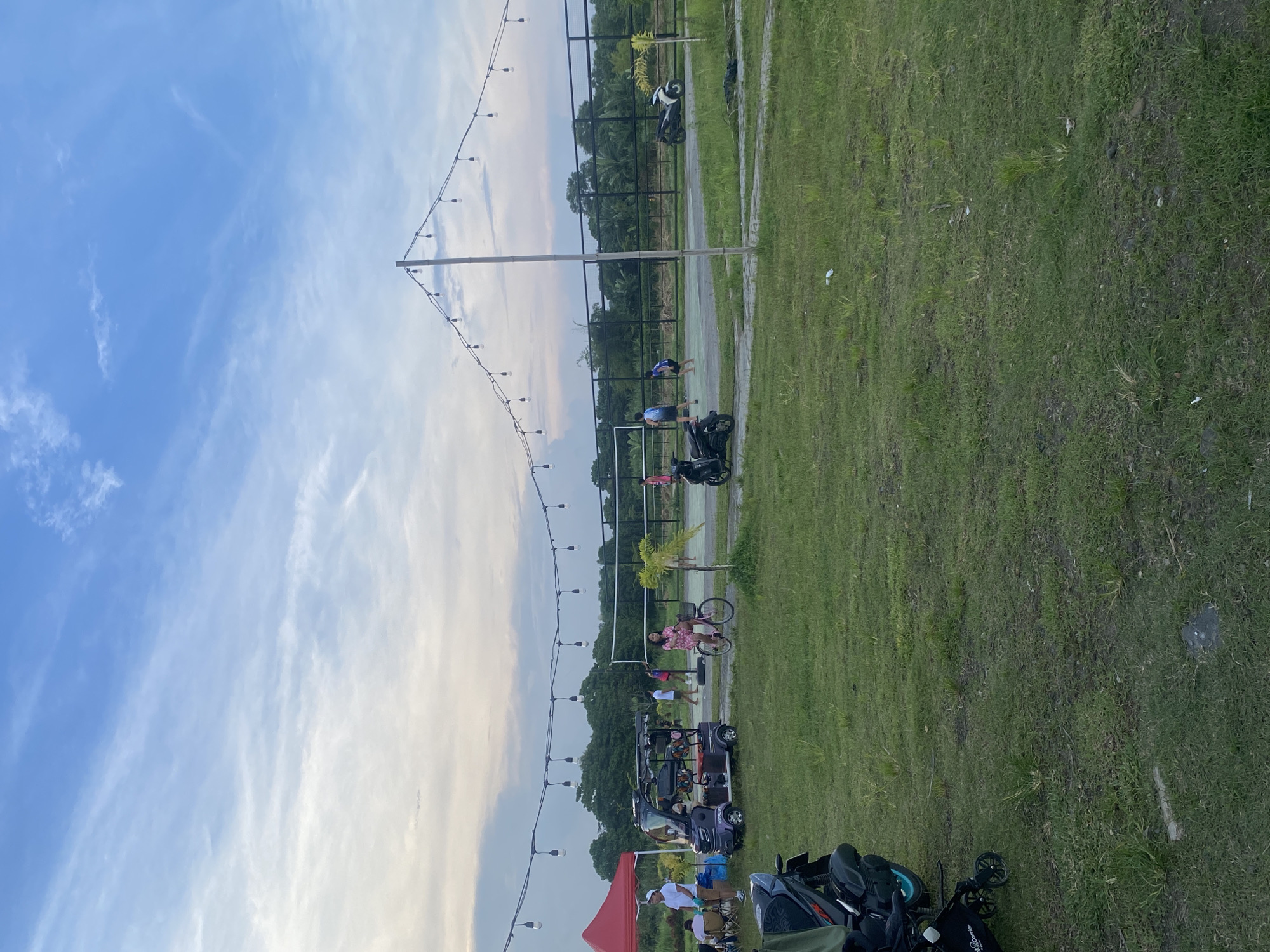 The image depicts an outdoor recreational area in Davao City, Philippines, under a clear, late afternoon or early evening sky with scattered white, wispy clouds. The scene is broadly divided into a vast expanse of green grass in the foreground and right, and a bustling activity zone with a blue sky overhead.
A prominent string of decorative light bulbs is suspended across the middle ground, supported by tall, slender poles, suggesting an ambiance for evening events. Behind these lights, a fenced sports court with a green playing surface is visible, where two individuals in blue tops are present. Nearby, a person in a pink patterned shirt stands next to a bicycle, and a white motor scooter is parked.
Closer to the foreground, a purple golf cart is parked, with several people gathered around it. A red canopy tent is set up, providing shade for a group of individuals seated on chairs. Multiple motorbikes and scooters are parked in this area, along with a baby stroller on which the brand "cybex" is faintly visible. The background features a line of lush trees, indicating a natural setting.
The image captures a casual gathering or leisure event, with people engaging in various outdoor activities or simply relaxing. The lighting suggests a time of day transitioning from afternoon to dusk, contributing to a relaxed and social atmosphere.
