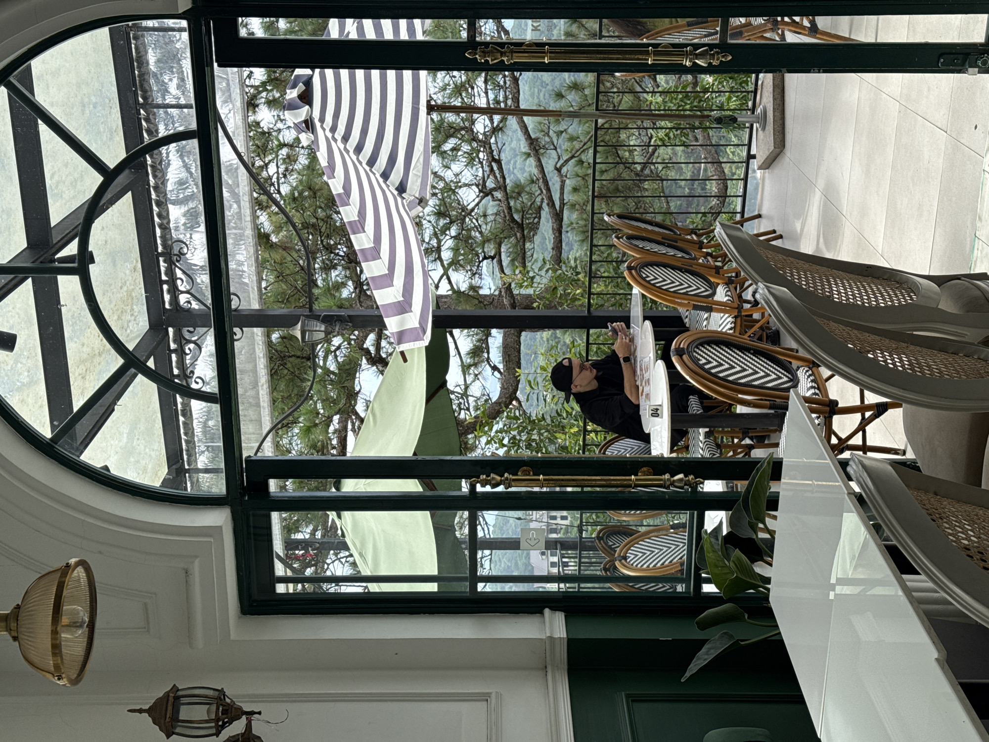 An overhead lamp and a wall sconce hang from the white ceiling of a room. The room has a large arched window with a glass roof. Through the window, an outdoor patio with tables and chairs is visible. A man in a black shirt and cap is sitting at one of the tables, looking at his phone. He is wearing glasses and a watch. A striped umbrella provides shade over another table on the patio. In the background, lush green trees and rolling hills can be seen. The patio appears to be part of a cafe or restaurant in Da Lat, Vietnam, a city known for its cool climate and beautiful scenery.