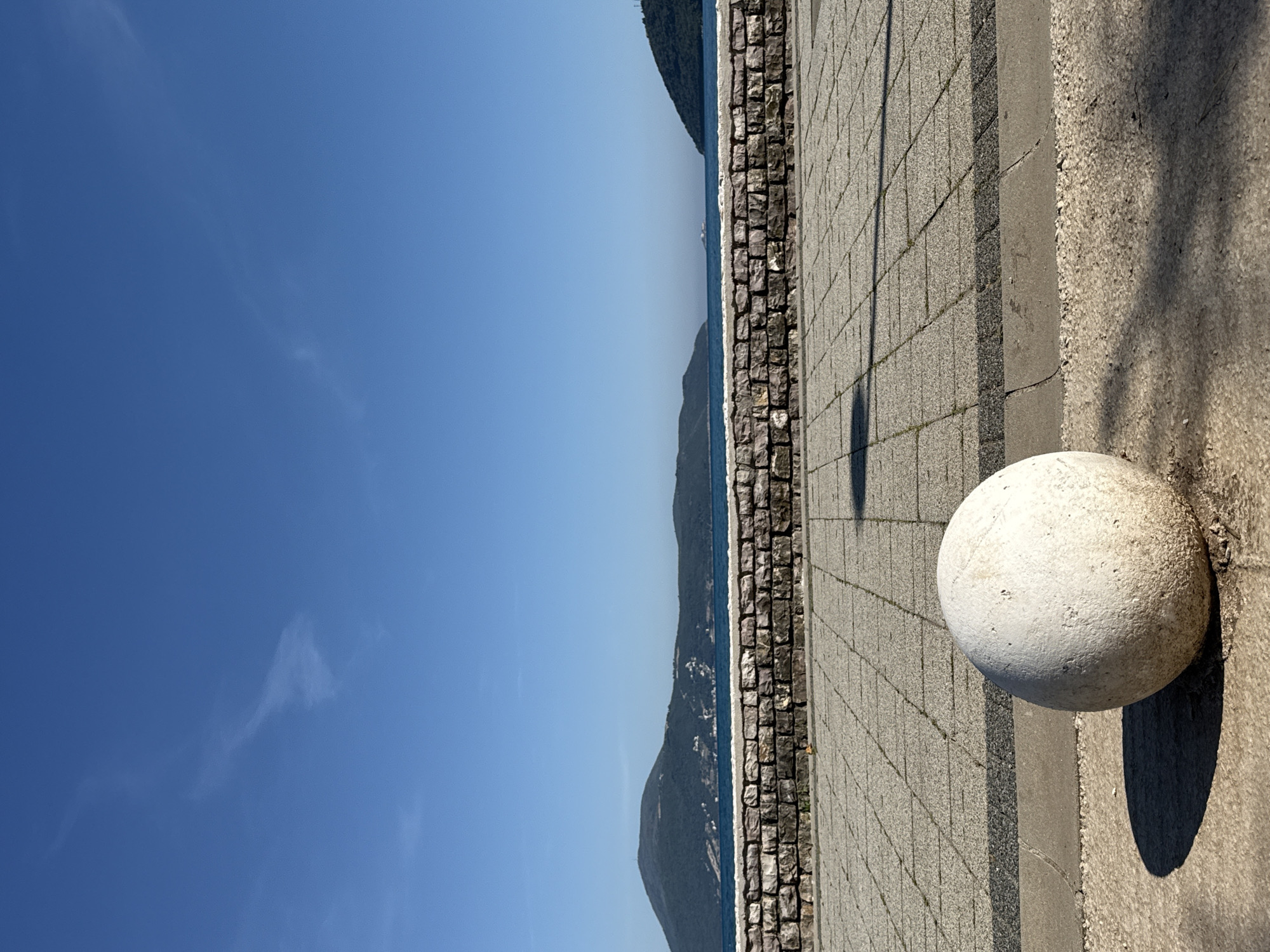 This image depicts a serene coastal scene in Igalo, Montenegro, under a bright, clear sky. In the foreground, on the right side, a large, light-colored spherical stone bollard sits on a textured, gravel-like ground surface. It casts a prominent, elongated oval shadow towards the lower left. Adjacent to this, a neatly paved promenade made of square grey tiles stretches horizontally across the image, separated by a dark grey curb.
To the left of the tiled walkway, a low stone wall, constructed from rugged, dark grey blocks, forms a boundary. Beyond this wall, a calm, deep blue body of water, likely the sea, extends into the distance. Across the water, a series of verdant, tree-covered hills or mountains rise, their silhouettes creating a gentle horizon line. The upper left portion of the frame is dominated by a vast expanse of clear, brilliant blue sky, with only a few very faint, wispy white clouds visible.
The long, distinct shadows cast by the bollard and along the promenade suggest strong, direct sunlight, indicating either an early morning or late afternoon setting. The weather is unmistakably sunny and clear. The scene exudes a peaceful and tranquil atmosphere. There are no people or discernible text visible in the image.