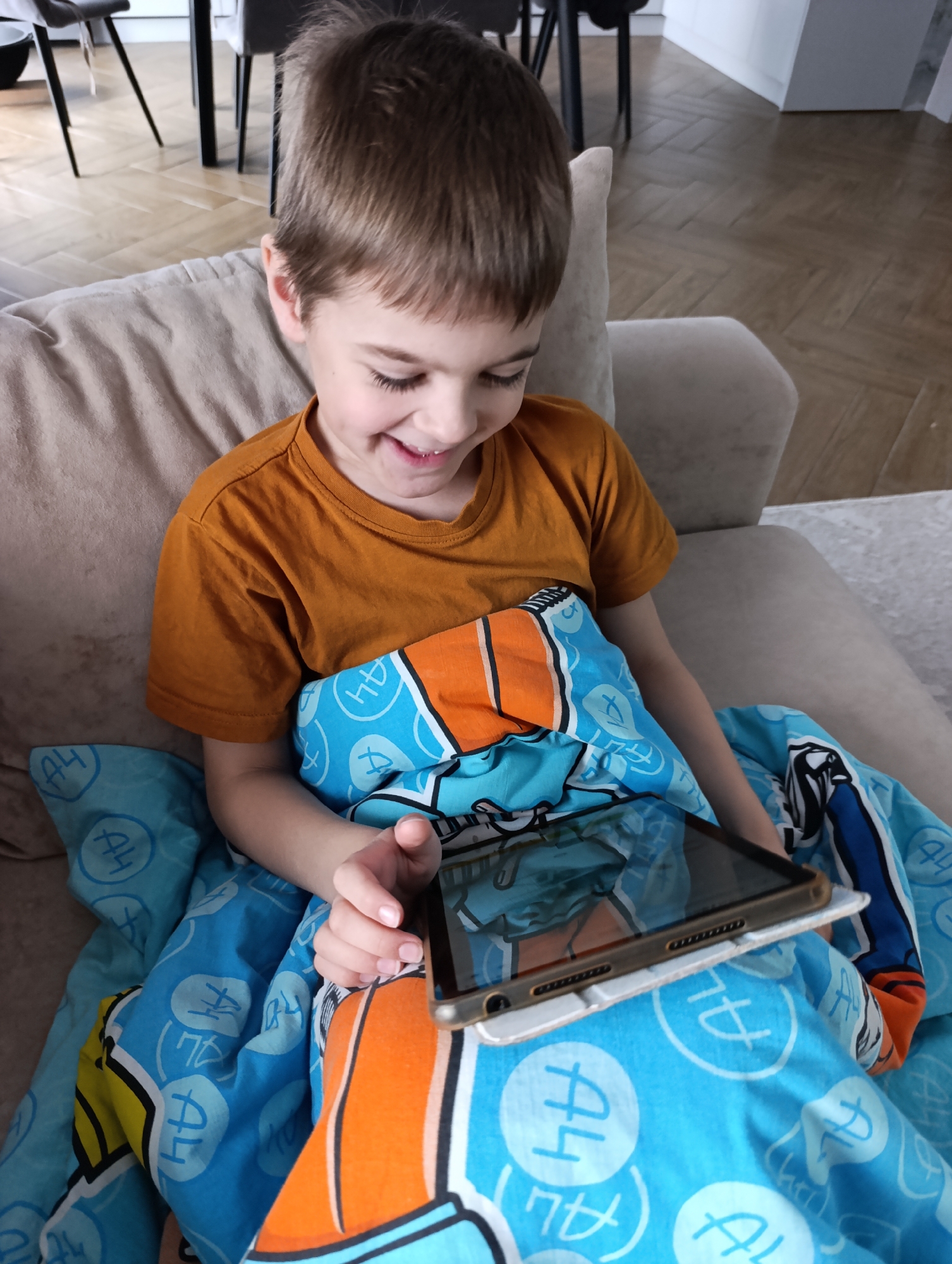 A young boy with short brown hair, estimated to be between 5 and 7 years old, is seated comfortably on a light beige sofa indoors. He is wearing an orange t-shirt and is covered by a blue blanket adorned with a repeating pattern of light blue outlined shapes and what appears to be the text "A4" or "AH," alongside other stylized symbols.
The boy is smiling broadly, with his head tilted down, fully engrossed in a tablet he holds with both hands. The tablet is resting on the blanket, and its screen reflects colorful content, likely a game or video, suggesting his cheerful and engaged expression. One of his fingers is positioned as if interacting with the touchscreen.
The setting appears to be a modern residential interior, possibly a living room or an open-plan space. A wooden floor with a herringbone pattern is visible in the background, along with the dark legs of chairs and a table, and hints of white cabinetry further in the distance. The lighting suggests it is daytime. The scene conveys a sense of relaxed enjoyment and focus on the digital content.