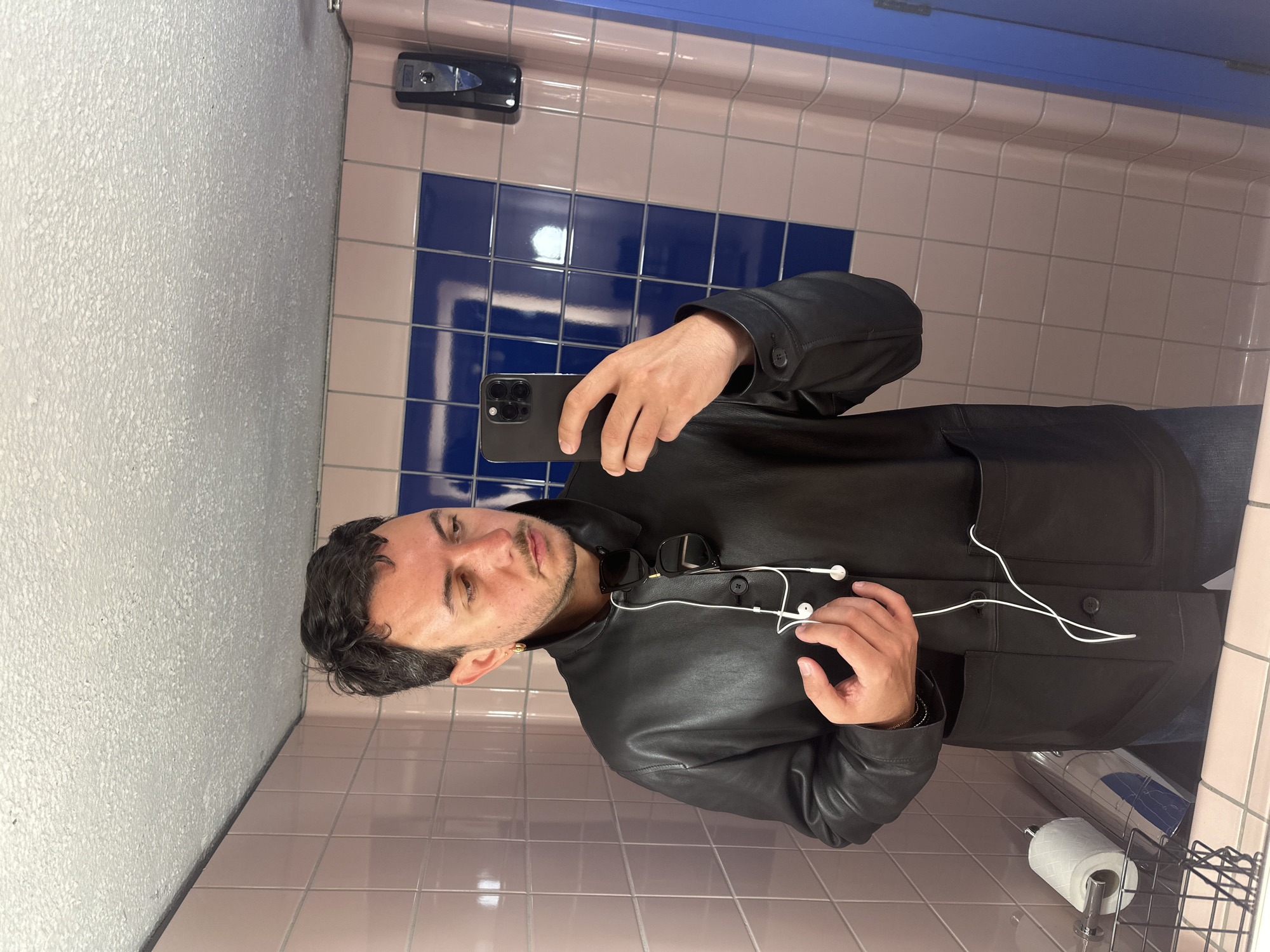 A man is depicted taking a selfie in what appears to be a public restroom in London, United Kingdom. The image is oriented sideways, but if viewed upright, the man is facing forward, looking directly at the camera. He has dark, wavy hair and a short beard or stubble, with a neutral to slightly serious expression. He wears a black leather jacket, with dark sunglasses hanging from the collar and white wired earphones draped around his neck. A small, gold-colored earring is visible in his left ear, and a gold-colored bracelet on his left wrist. He holds a dark-colored smartphone with a visible triple-camera setup in his right hand, positioned to capture the selfie.
The background consists of light pink tiled walls with a pattern of darker blue tiles arranged in a grid-like fashion, specifically seen as horizontal bands and some square patterns. Above the tiled section, there's a blue-painted strip and a textured white ceiling. To the right of the man, a chrome toilet paper holder with a roll of white toilet paper is visible. Further up on the wall, a black dispenser (possibly for soap or hand sanitizer) is mounted. Reflections, possibly from a toilet or a metallic bin, can be seen on the lower right side of the image. No visible text is present in the scene. The overall lighting suggests an indoor setting with artificial illumination.