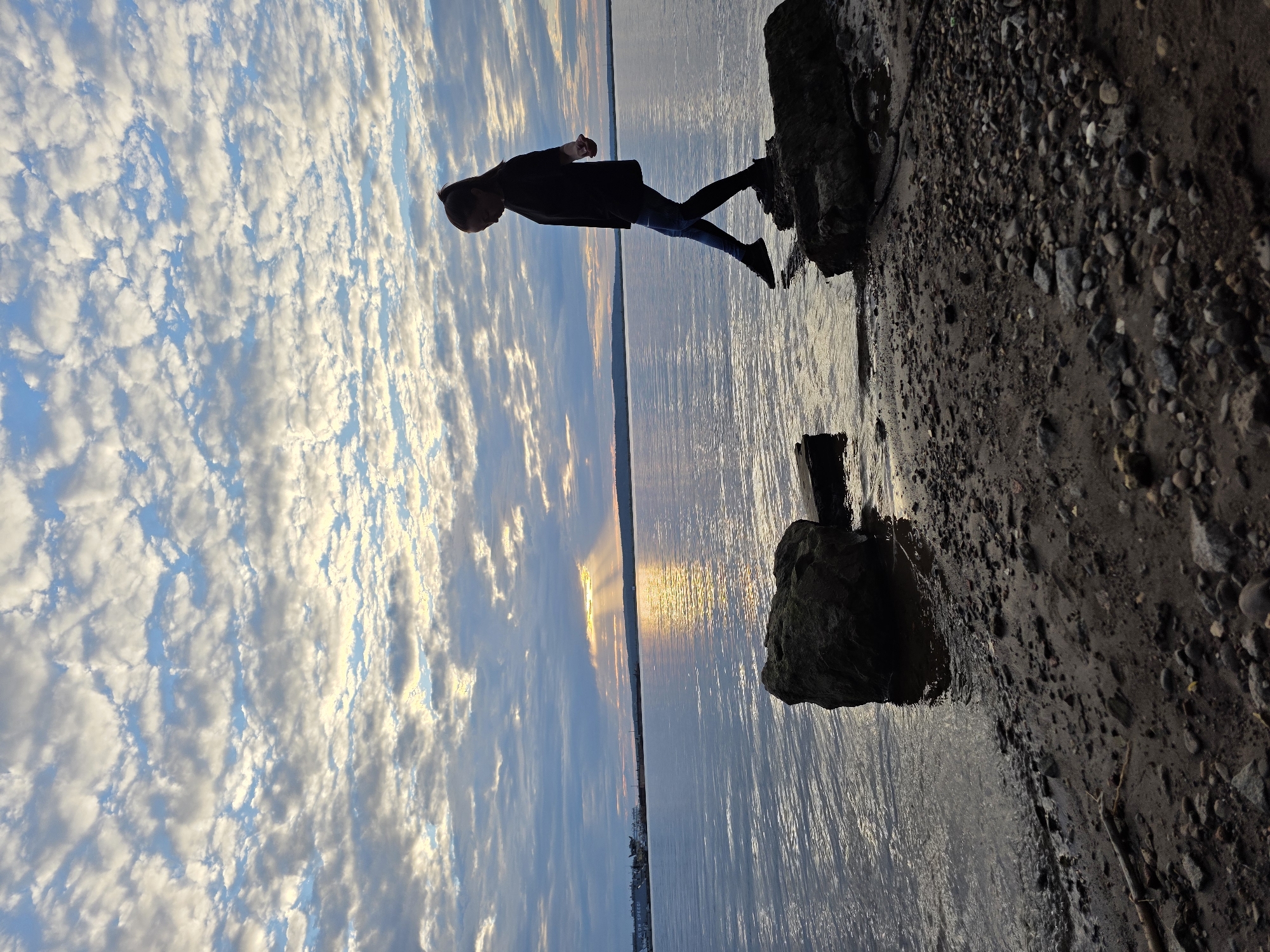 A full-frame, eye-level shot captures a woman silhouetted against a sunset over a body of water. She is positioned on the right side of the frame, mid-stride, stepping from one rock to another near the shore. The woman is wearing dark clothing and has her hair pulled back in a ponytail. The sky is filled with a dramatic display of clouds illuminated by the setting sun, creating rays of light that break through. The water's surface is rippled, reflecting the colors of the sky. The foreground consists of a rocky and sandy shore. In the distance, across the water, a faint outline of a city or town is visible on the horizon. This scene is located on the Kama River embankment in Chaikovsky, Russia.