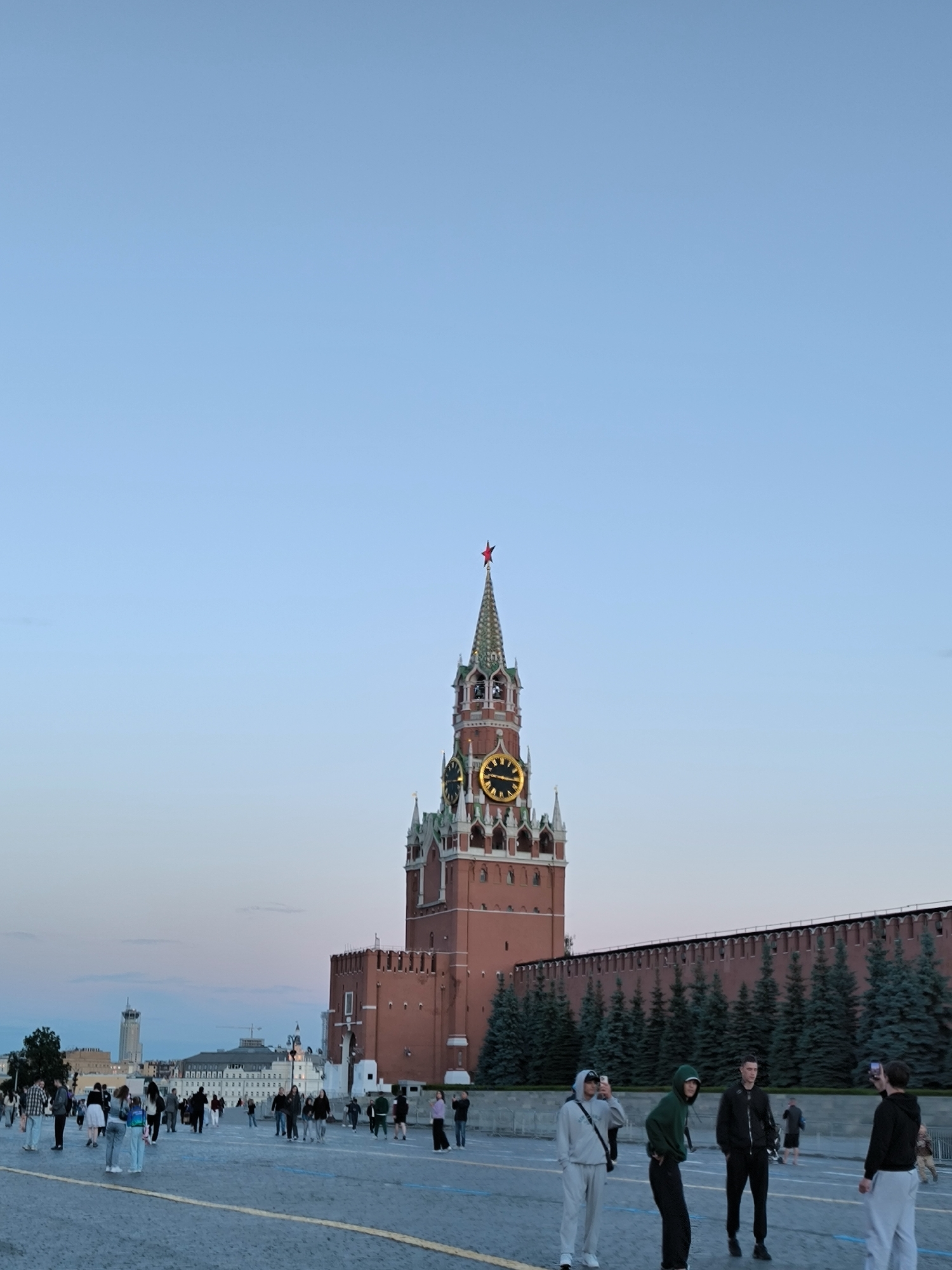 This media file captures a view of the Spasskaya Tower in Moscow, Russia. The tower, a prominent landmark, features a red star at its apex and a large clock with illuminated golden hands and numerals, suggesting it is either dusk or dawn. The sky is a soft blue, transitioning to a pale pink and orange hue closer to the horizon, indicating the twilight hours.
In the foreground, a paved plaza is populated by a diverse group of people casually walking and conversing. Some are taking photographs with their phones, engaged in typical tourist activities. Their attire is casual, suggesting a relaxed atmosphere. Behind the people and to the right, a section of the red brick Kremlin wall, topped with a row of dark green evergreen trees, forms a backdrop. Further in the distance, beyond the plaza, are several buildings, including a tall, modern tower and other structures that contribute to the urban landscape of Moscow. The overall scene depicts a popular public space at a transitional time of day, filled with people enjoying the surroundings. There is no visible text in the image.