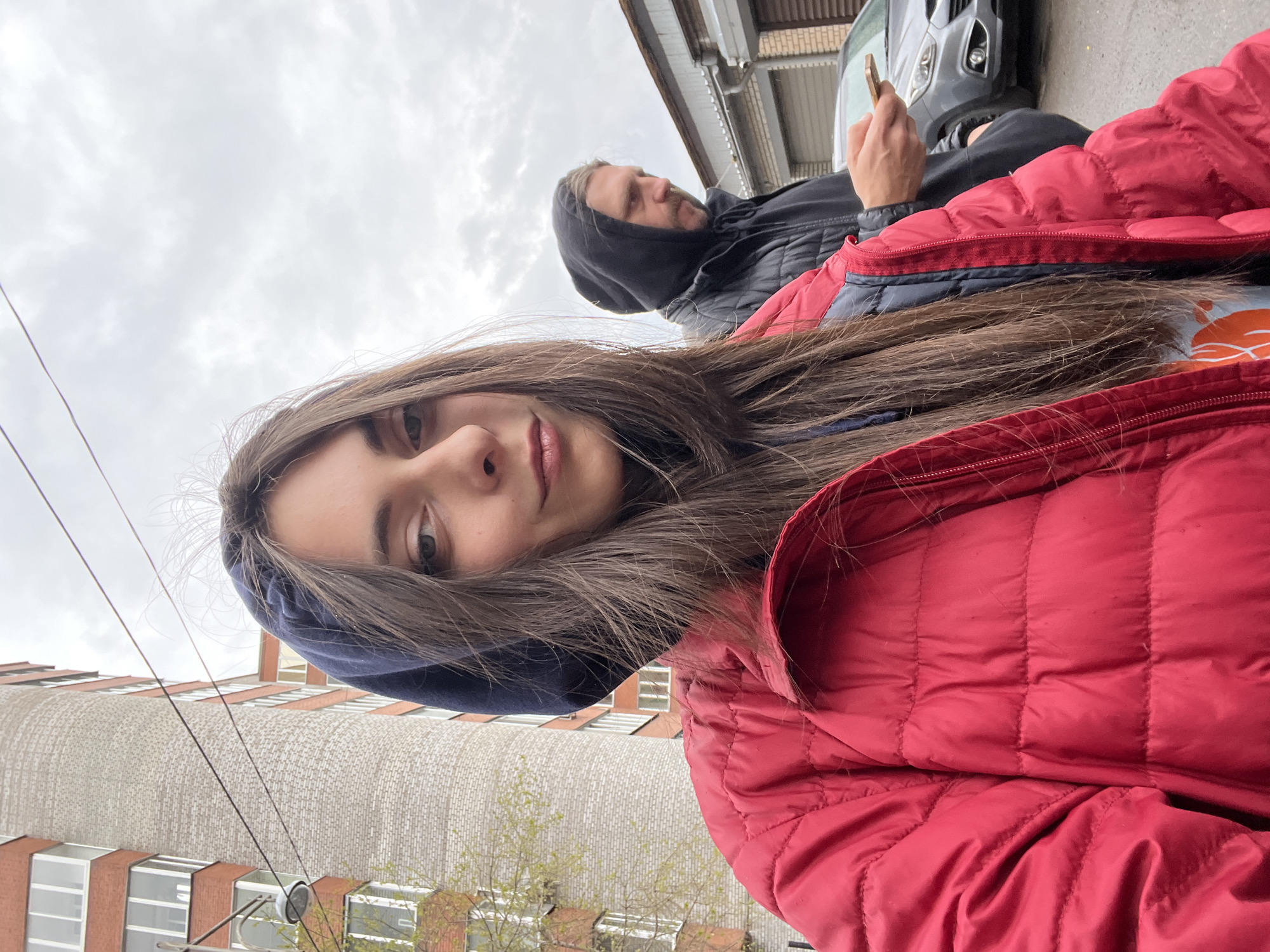 A selfie photograph depicts a young woman in a red puffer jacket with the hood up. Her hair is long and brown. In the background is a man with a hooded black jacket and a mobile phone in his hand. A gray car is partially visible behind him, and a building with windows is in the background to the left. The sky is overcast. Based on architectural style and other details, it is likely that the photo was taken in a city in Russia, possibly Krasnoyarsk.