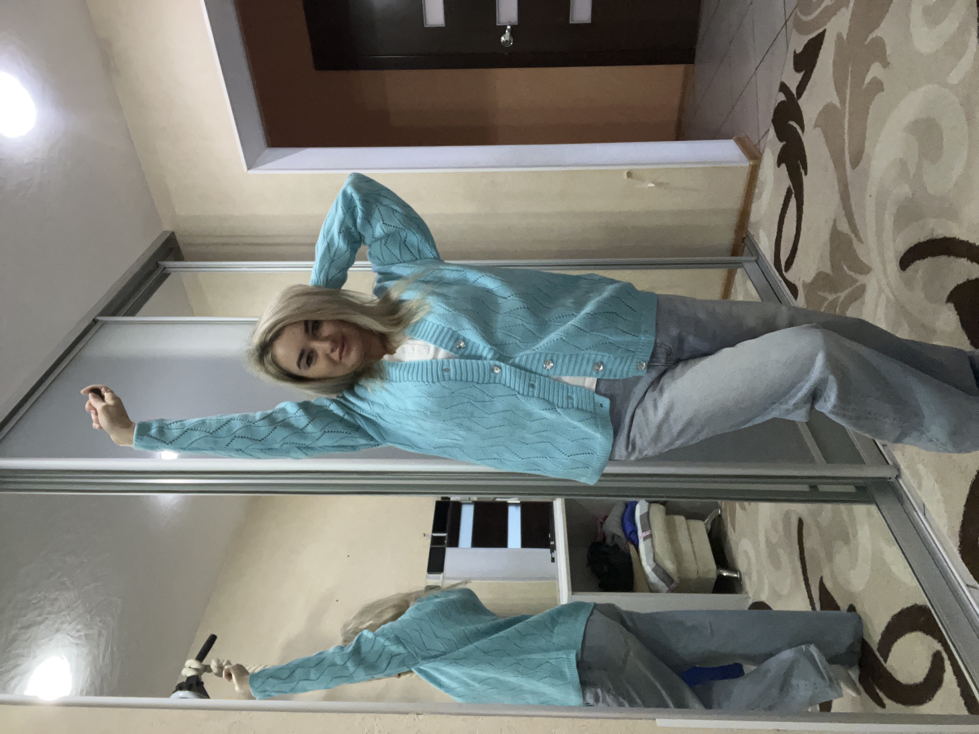 A woman with blonde hair is posing in front of a mirrored sliding door closet. She is wearing a light blue cardigan and grey jeans. The closet is in a room with beige walls and a patterned rug. Reflected in the mirror is a partial view of the room's interior, including shelves and possibly other clothing items within the closet. The image was taken in a location in the city of Chaikovsky, Russia.