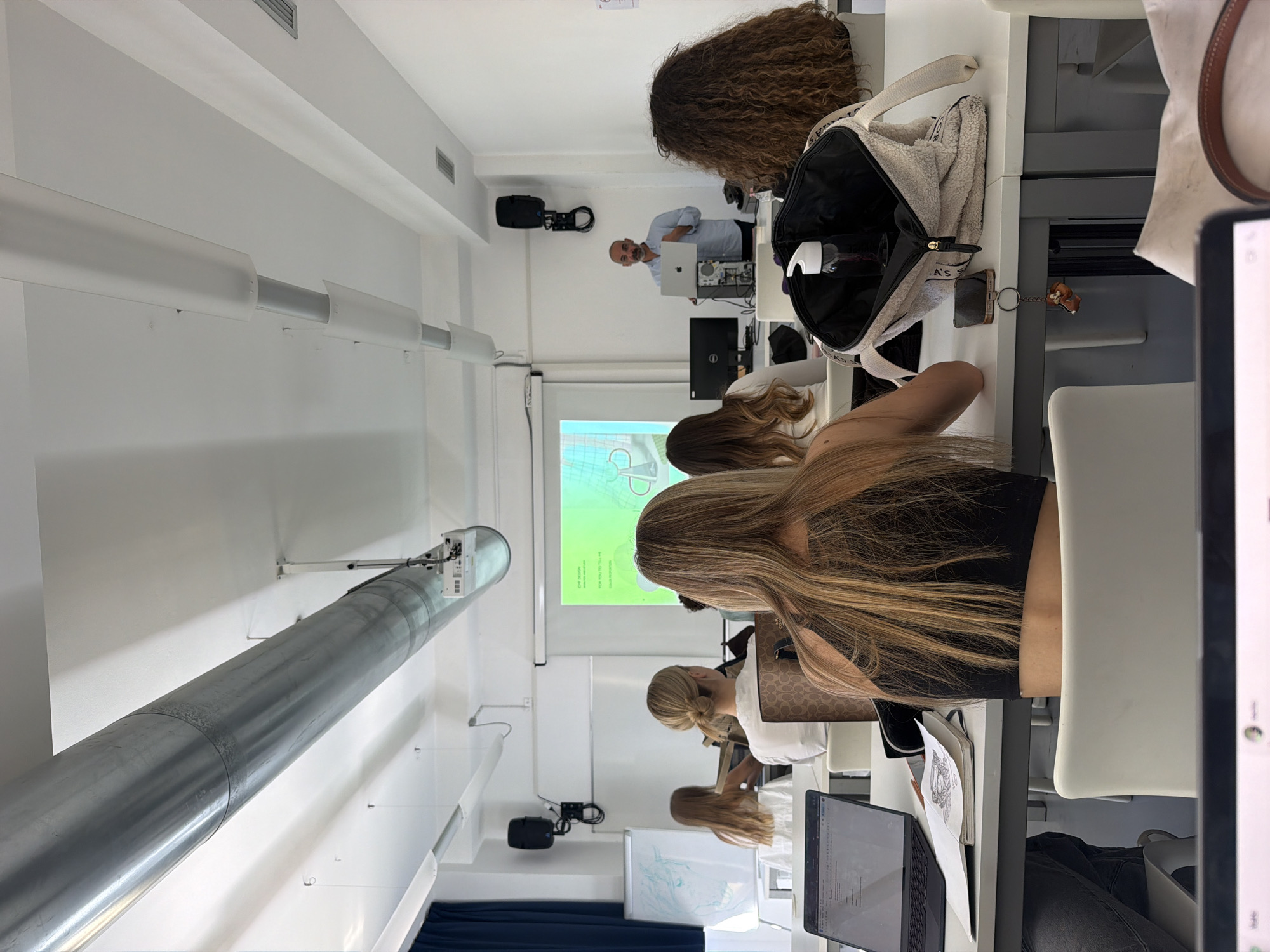 The image depicts a classroom scene, likely in Milan, Italy. A middle-aged, light-skinned male instructor is visible at the front of the class, using a laptop. Several students are seated at desks, facing the front. The hair colors of the students vary, with blondes, brunettes, and one student with curly brown hair. Many students have laptops open in front of them. There's a projector screen with what appears to be an architectural diagram visible. Overhead, a large metallic ventilation duct runs the length of the room. Personal items like bags and water bottles are visible on the desks. The room is generally white-walled and appears to be well-lit. There is a blue curtain at the back of the room.