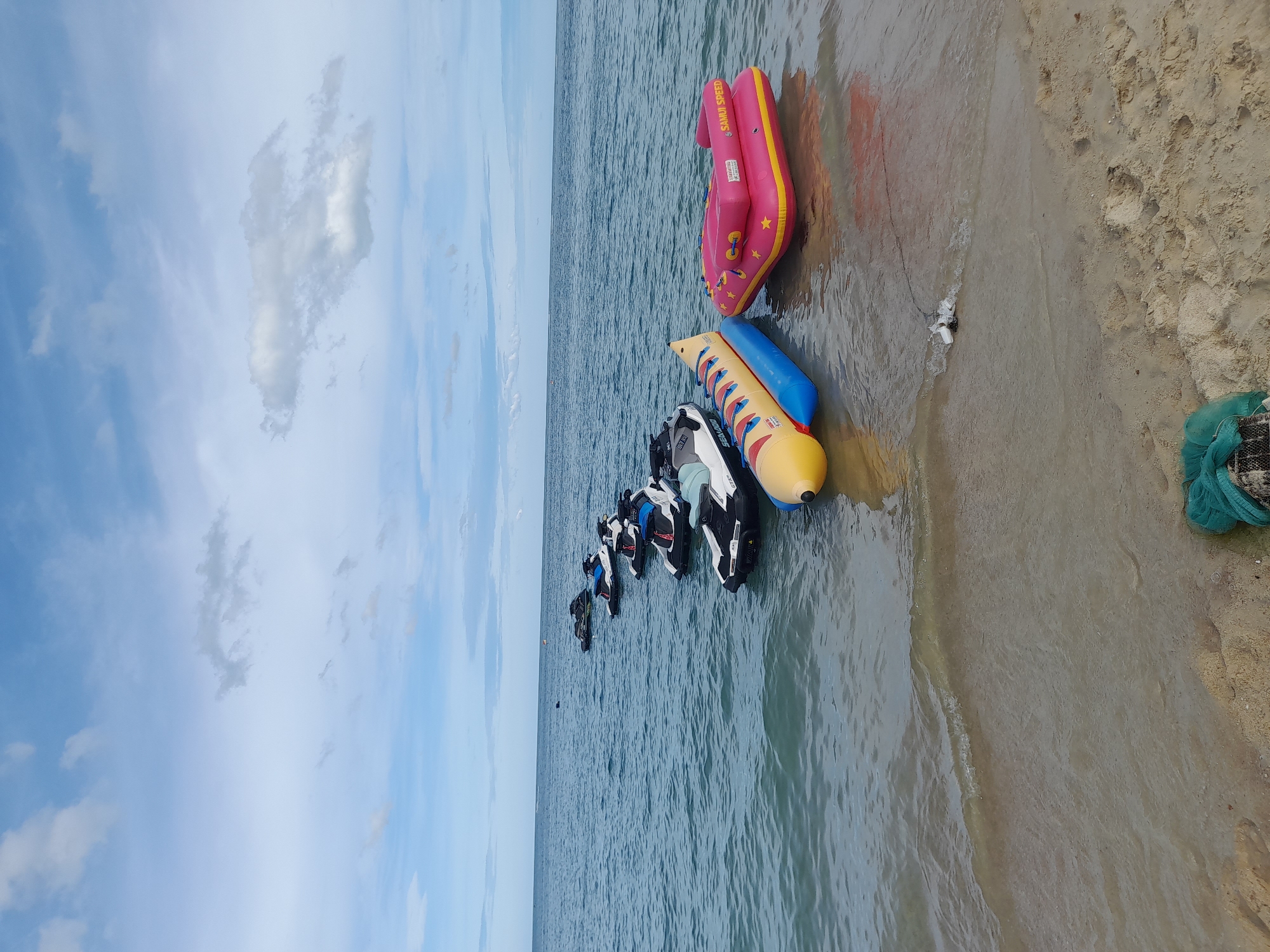 In the ocean off the coast of an unknown location, a line of jet skis is anchored in the water. In the foreground, closer to the sandy shore, a yellow banana boat and a pink inflatable raft are partially submerged in the shallow water. The sky above is a mix of blue and white clouds. The image is a wide shot, capturing the expanse of the sea and the beach.