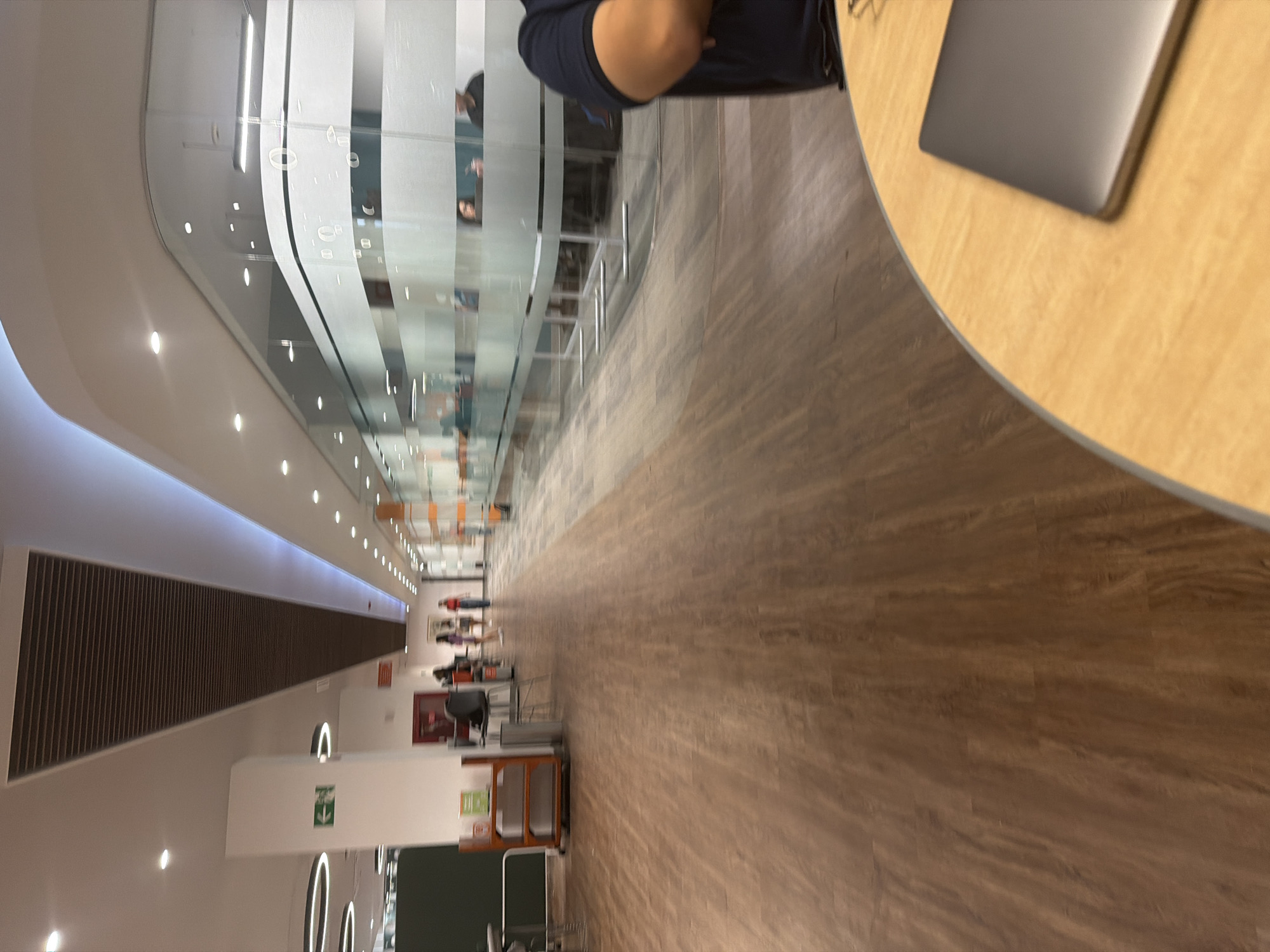 The image depicts a long, well-lit hallway in a modern building, possibly an academic or office space in San Andrés Cholula, Mexico. The hallway is predominantly brown with wooden flooring and a large, curved glass wall on the left, behind which are office spaces with people visible inside. Light fixtures line the ceiling. On the right, near the foreground, is a round, wooden table with a closed gray laptop on top. Further down the hallway on the left, there is a ventilation system, chairs, and what seems to be an exit sign and a chalkboard. There are a few people walking at the far end of the hallway. A person is partially visible on the upper-right of the image, near the table.