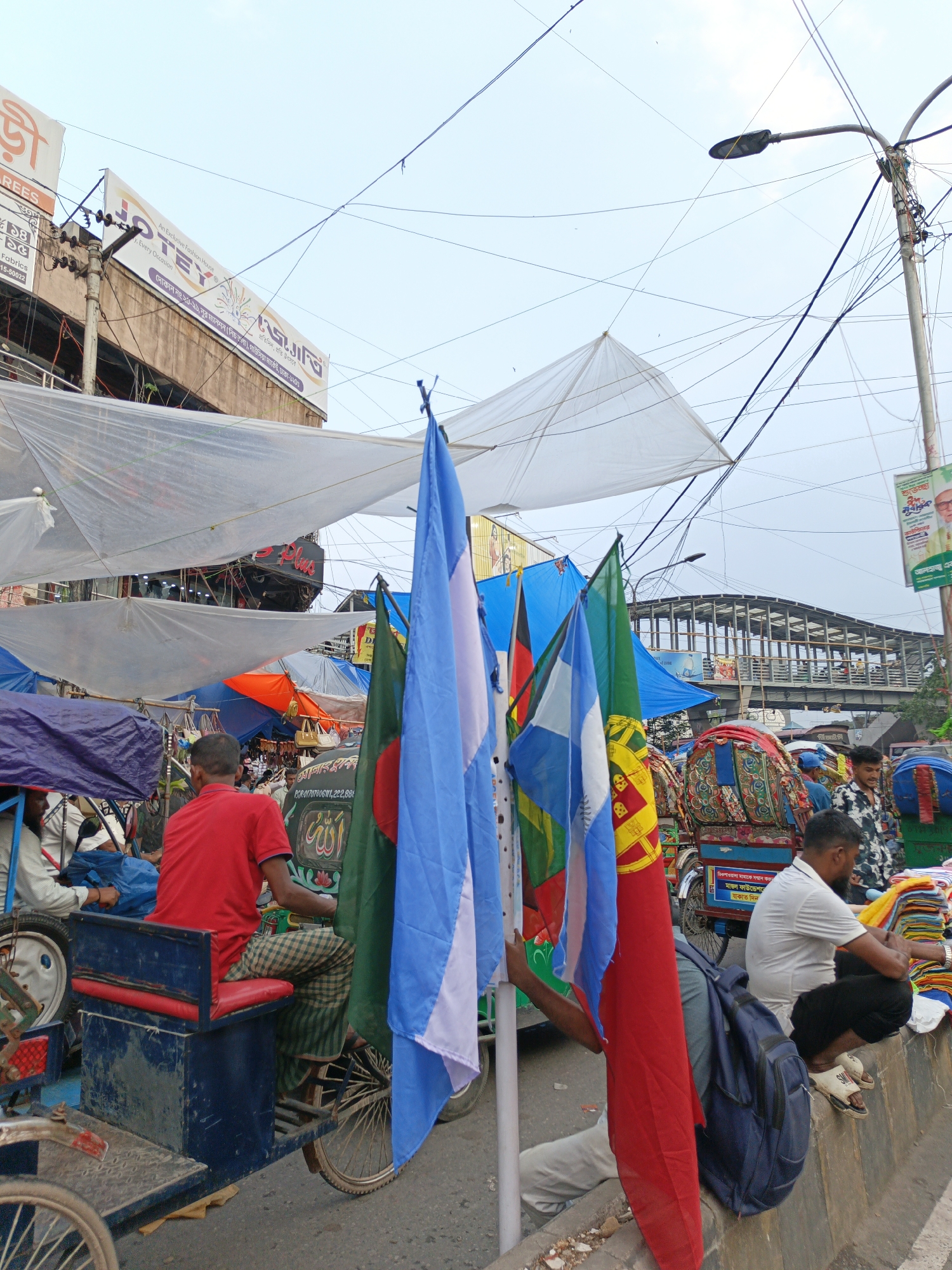 The image captures a street scene, likely a market, in an urban environment. The sky is overcast, and a network of electrical wires crisscrosses the upper portion of the frame, indicating a densely populated area. Several makeshift awnings made of translucent white material are suspended above, creating shade. In the foreground, a person in a red shirt sits on a blue rickshaw. Flags of various countries, including Bangladesh, are prominently displayed, suggesting a potential celebration or event. Several other individuals are visible in the background, some engaged in activities related to commerce, like stacking goods or tending to rickshaws, while others are simply present in the environment. The presence of multiple rickshaws, some elaborately decorated, points to a location where these vehicles are a common mode of transportation. The overall impression is one of a busy and vibrant street market, with a mix of commercial activity and everyday life.