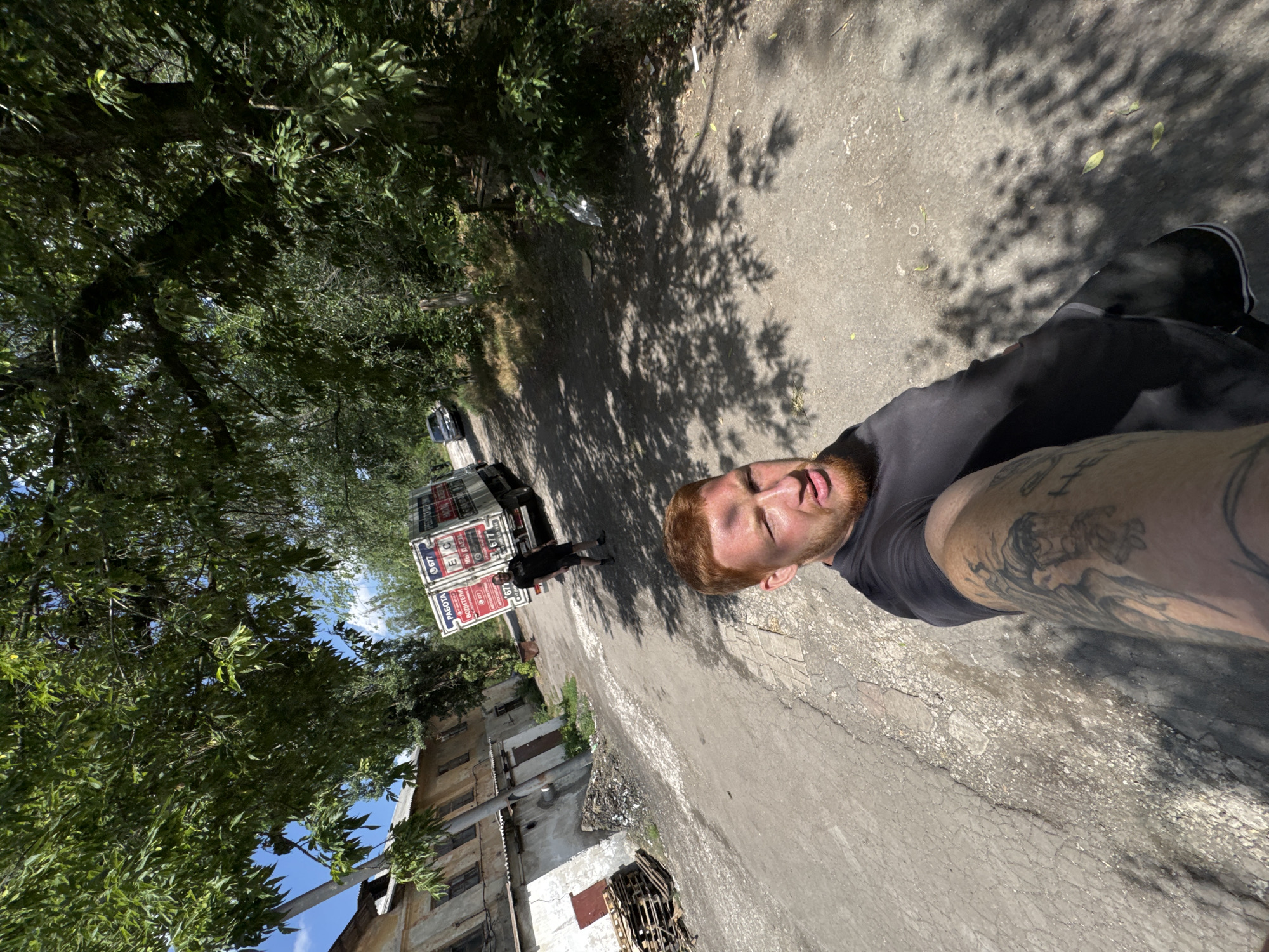 An outdoor, eye-level, wide shot captures a man with red hair and a beard taking a selfie with his left arm extended. He is wearing a black t-shirt and black shorts, with visible tattoos on his left arm. A bruise is visible on his forehead. In the background, a truck with advertisements on its side is parked on an unpaved road, with a dark sedan parked further down the road. An old, light-colored building with a damaged facade stands to the left of the road, with a pile of wooden pallets and debris in front of it. Lush green trees with full foliage line the right side of the road, casting dappled shadows on the ground. The sky is visible through the tree canopy, with patches of blue and white clouds. The scene appears to be in an industrial or semi-abandoned area, with an overall atmosphere of disrepair.